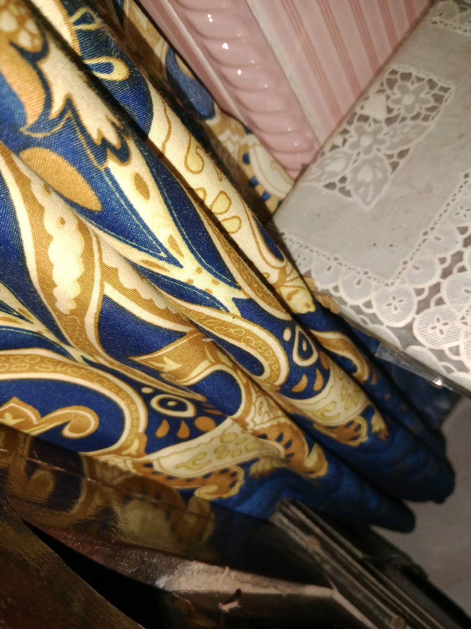 A close-up, low-angle shot captures folded fabric with a blue and gold paisley pattern, layered over other textiles. To the right, a white lace doily with floral motifs is visible, and behind that, pink fabric with subtle stripes. The lower portion of the image reveals a dark, reflective surface, possibly wood or metal, with indistinct objects. The scene is indoors, with artificial lighting. There are no people present. The setting is too generic to determine a specific location within Ringinrejo, Indonesia.