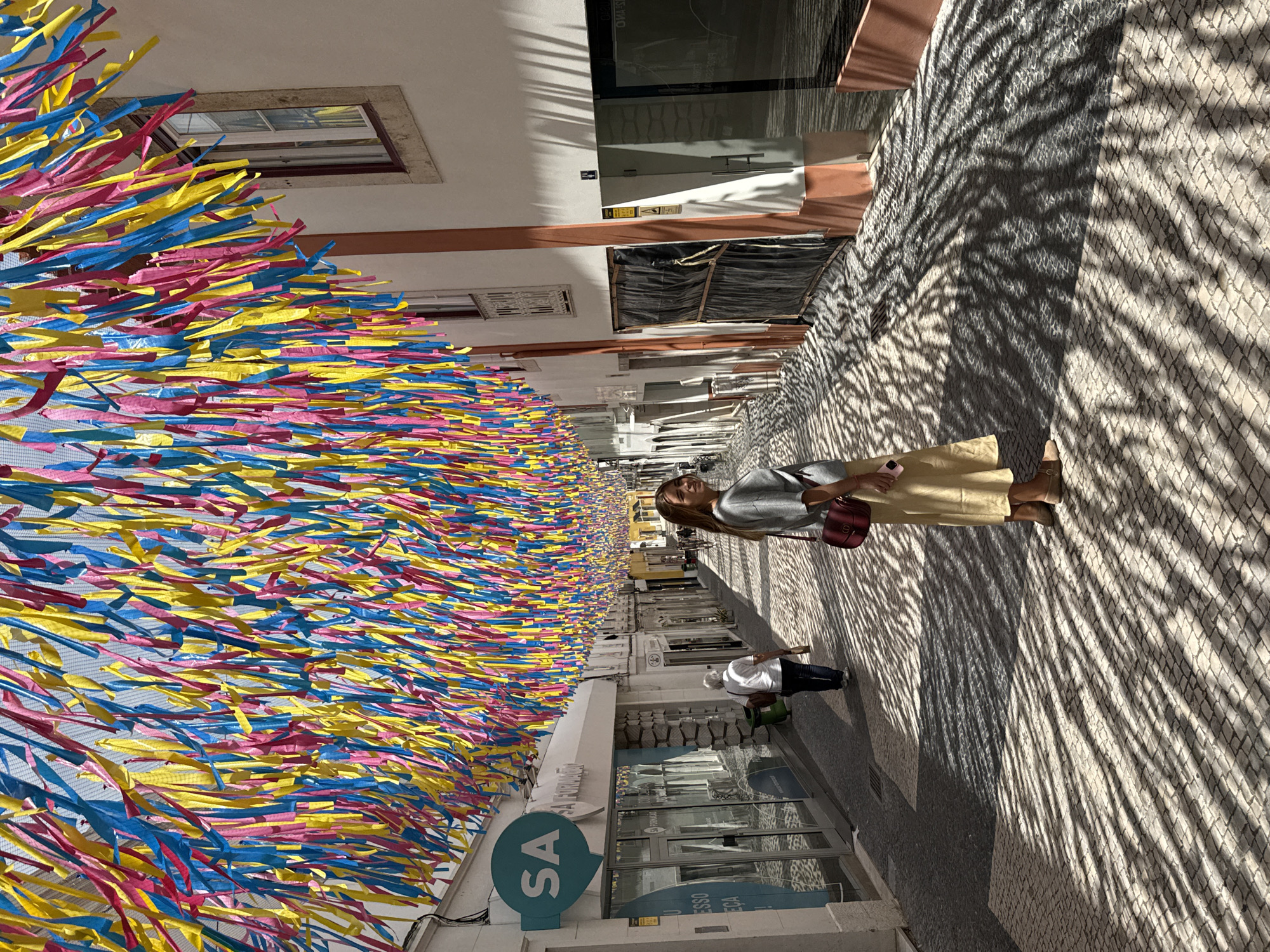 A young woman with blonde hair, wearing a grey sweater, a beige skirt, and a red purse, stands on a cobblestone street in Torres Vedras, Portugal. The street is decorated with colorful streamers, creating a festive atmosphere. Sunlight streams through the streamers, casting striped shadows on the ground. The woman is looking back over her shoulder, her expression relaxed. In the background, other people are walking down the street, and shops line the sides of the road. The buildings are white with orange trim, and windows reflect the sky. The street appears to be a pedestrian area, with no cars visible. The overall impression is one of a vibrant and lively urban environment.