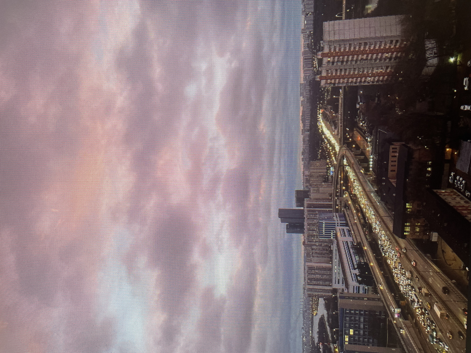 The image depicts a twilight cityscape with a prominent sky filled with pink and purple clouds. In the mid-ground, a multi-lane highway is congested with traffic, characterized by numerous headlights and taillights. Several large apartment buildings, some with illuminated windows, are visible on either side of the highway and in the distance. A body of water, likely a river or canal, can be seen to the left, bordered by more buildings. In the far distance, a dense urban landscape stretches to the horizon, with illuminated streetlights and buildings indicating a populated area. Tall, dark skyscrapers are clustered together in the middle distance. The overall scene conveys a busy urban environment transitioning from day to night under a dramatic sky. The provided image is not of Sinyavino, Russia.