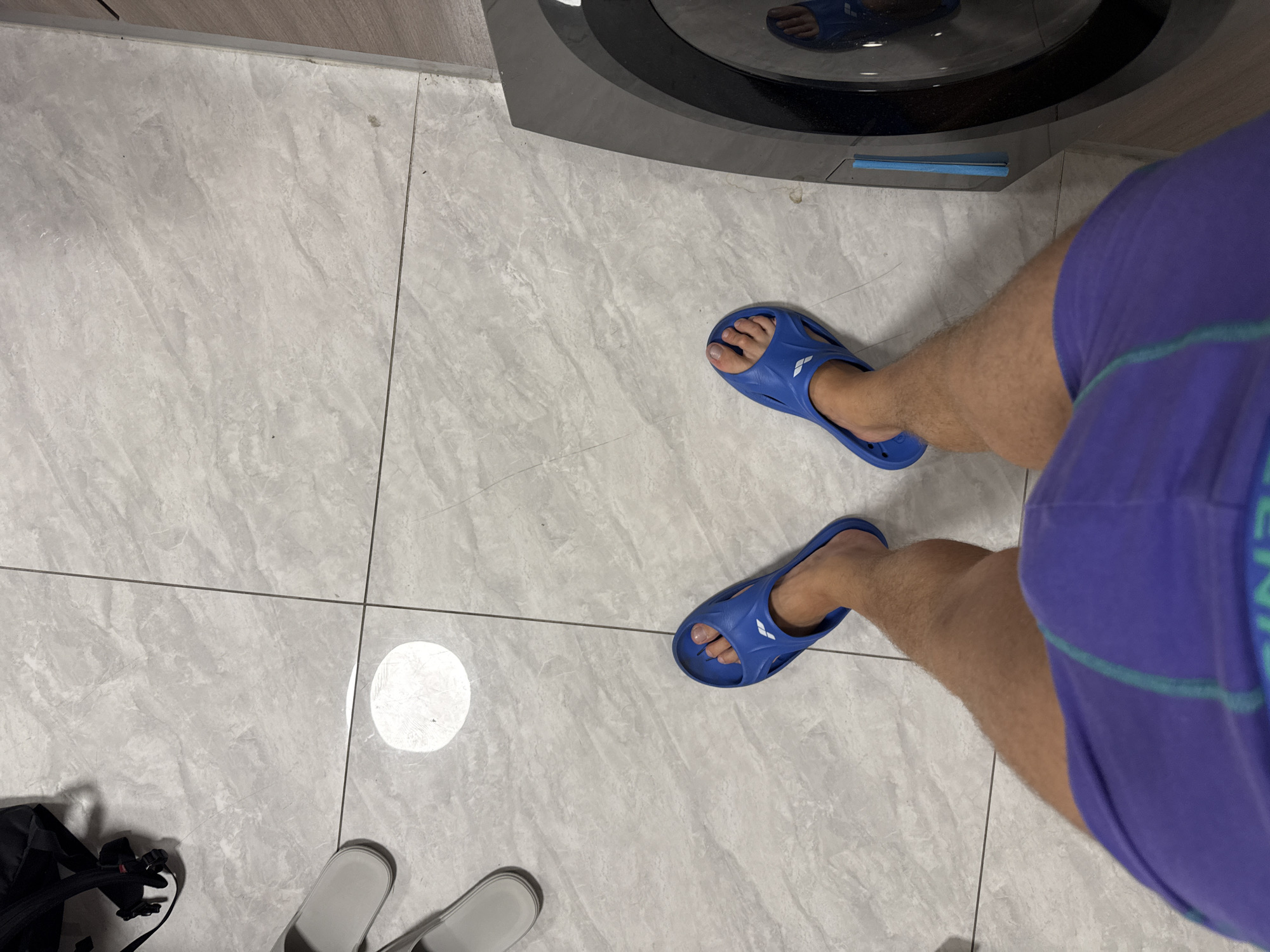 A person's legs, wearing purple athletic shorts and blue flip-flops, are shown from a top-down perspective on a tiled floor in what appears to be a laundry room, based on the presence of a washing machine on the right side of the image. The floor is made of light grey marble-patterned tiles, with dark grout lines. A black backpack is visible in the upper left corner of the image, and two grey slippers are on the floor near it. A circular reflection, possibly from a light source, is present on the tiles in the upper left quadrant. The washing machine has a circular glass door and a metallic gray exterior. The person's legs are tanned, and body hair is visible on the lower legs. The purple shorts have a light green stripe along the side. The blue flip-flops are the brand Arena. The image is likely taken indoors in a residential or commercial setting in Tianhe District, China.