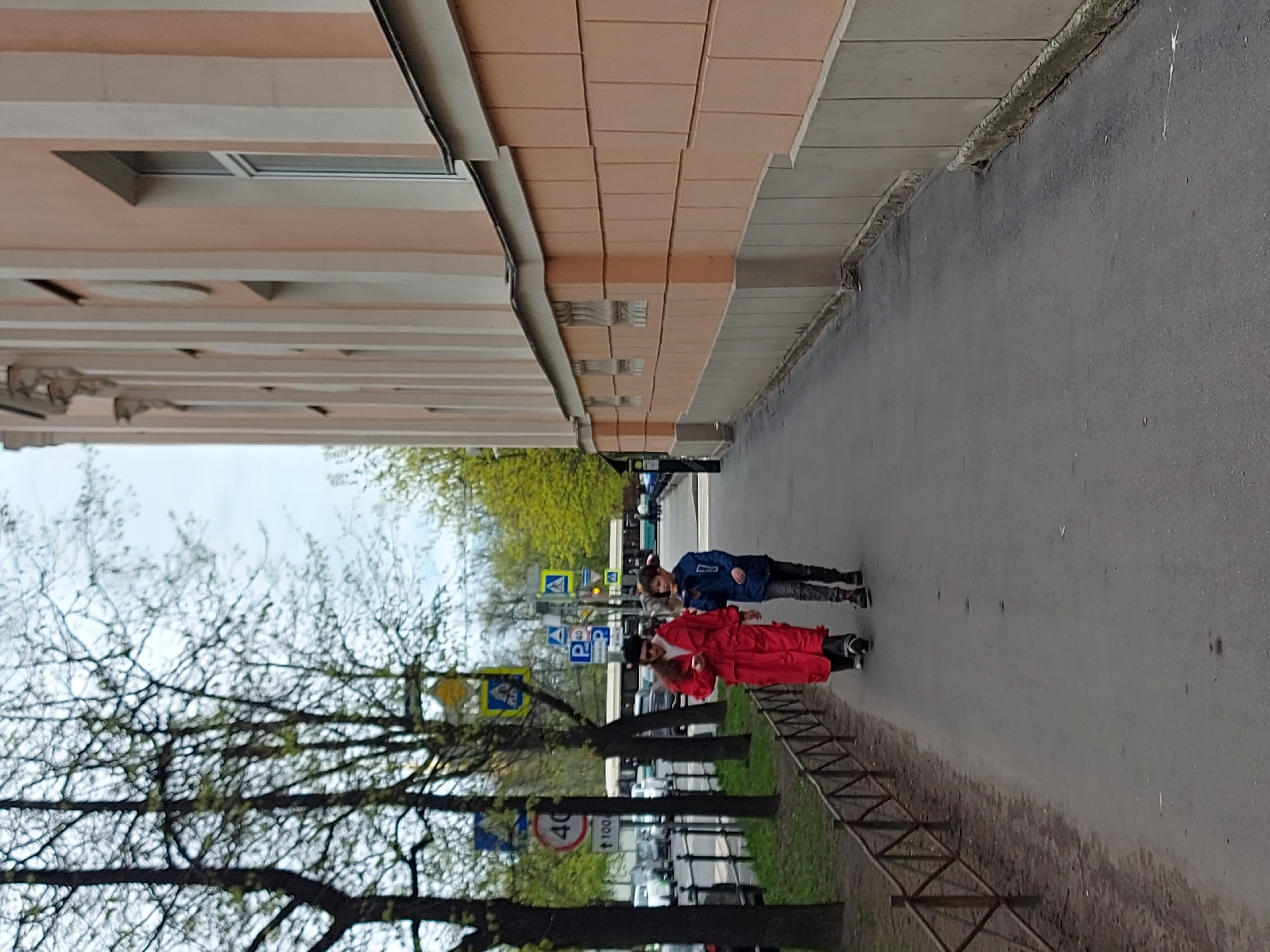 The image shows two people walking on a sidewalk alongside a building in Saint Petersburg, Russia. One person is wearing a red robe-like garment, while the other is wearing a blue jacket. The building is peach-colored, and trees are visible on the left side of the sidewalk. Street signs are also visible. Cars are parked nearby. The sky is overcast.