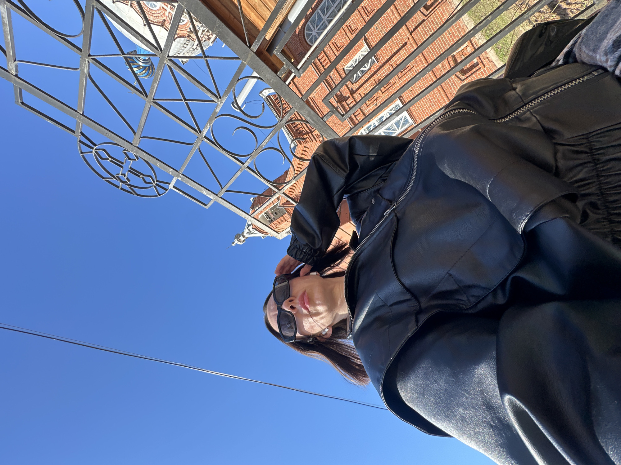 A low-angle view captures a woman in sunglasses and a black leather jacket, with a building and an ornate metal fence overhead against a clear blue sky. The building, partially obscured by the fence, is likely the Holy Transfiguration Cathedral in Votkinsk, Russia, given its brick construction and visible architectural details. Powerlines are also visible against the sky.