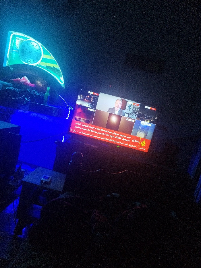 The image shows a dimly lit room with a television screen displaying a news broadcast. The news channel logo "Al Jazeera" is visible. The text on the screen appears to be in Arabic. In the top left of the image, there's a lit clock with a transparent face. A small table with an object on it is positioned to the left of the screen. An indiscernible object or person appears to be seated in front of the television screen. The environment is dark. The location in the city is Unknown, Unknown.