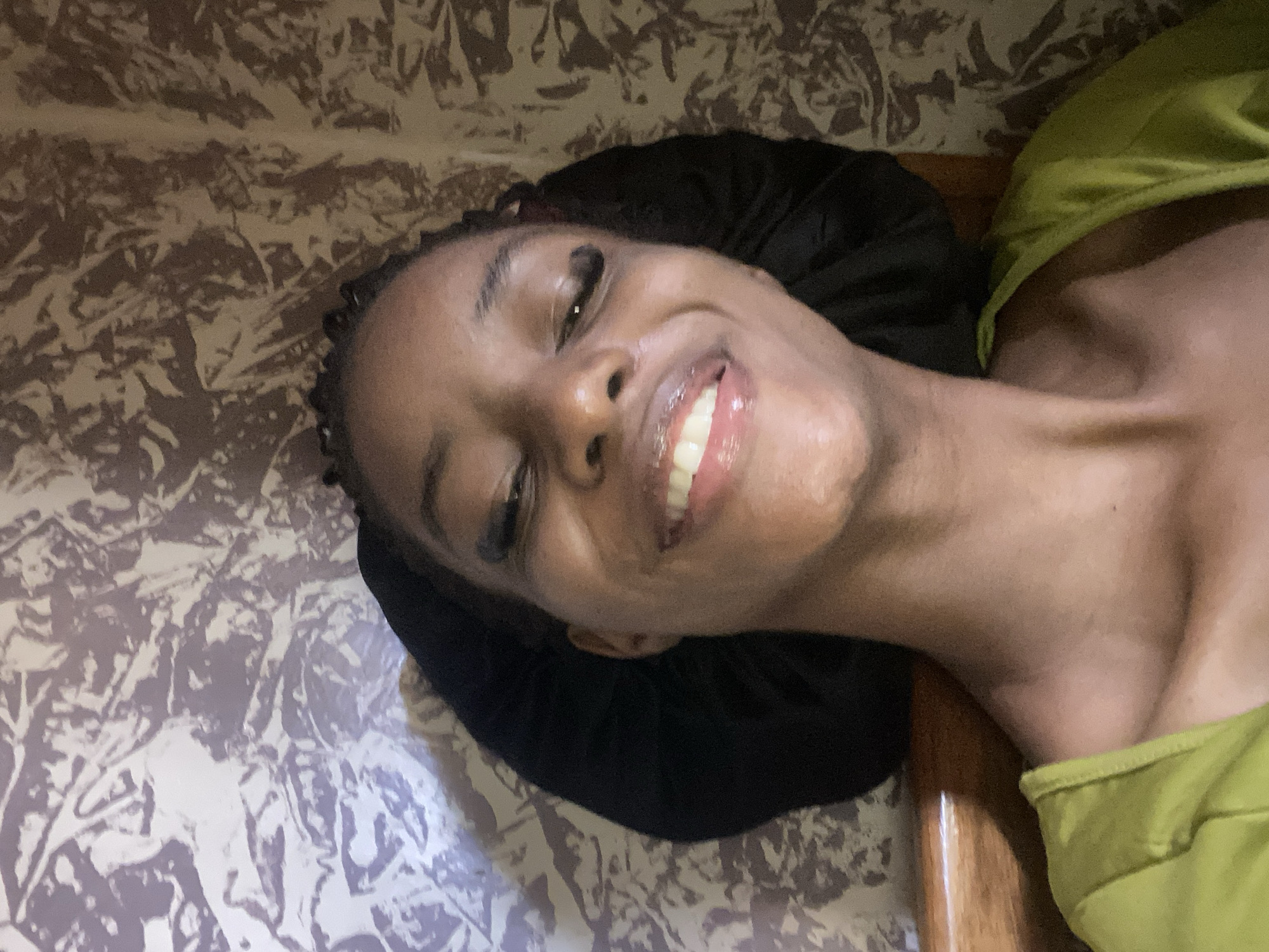 A close-up, upside-down selfie shows a young Black person smiling broadly, with their head tilted to the left. They are wearing a black bonnet and a bright green garment, possibly a top. The background features a textured wallpaper in shades of brown and beige, with a pattern that appears abstract or floral. A wooden surface is visible on the right side of the image. The lighting suggests an indoor setting, possibly at night or in a dimly lit room. The subject's expression is joyful and relaxed. No text is visible in the image.