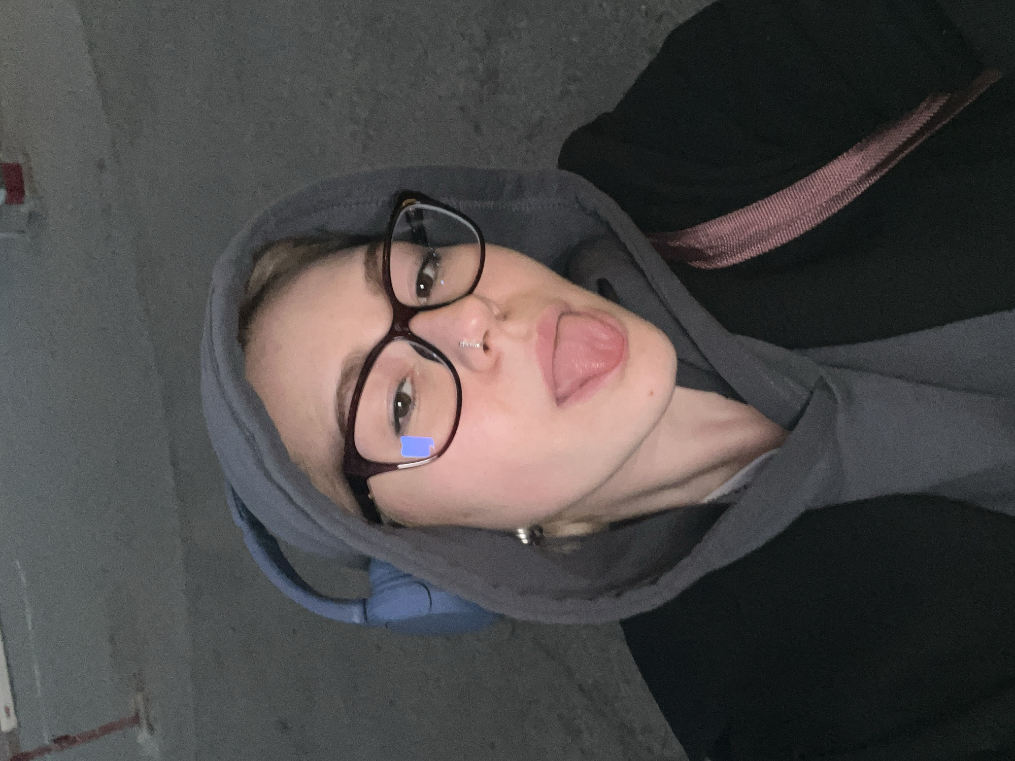 A rotated indoor selfie features a young individual, likely female, wearing a grey hooded sweatshirt, dark-framed glasses, and blue over-ear headphones. The person's head is tilted, and they look directly at the camera with a playful expression, their tongue slightly sticking out. A silver nose ring is visible on their left nostril, and a small silver earring adorns their left earlobe. A prominent blue rectangular reflection, possibly from a screen, is visible in the left lens of their glasses.
Underneath the grey hoodie, a black garment is visible, along with a dark pink or maroon ribbed strap, likely from a bag or another accessory, draped over their right shoulder. The background is a uniform, textured grey wall, possibly concrete, indicating an indoor or semi-enclosed utilitarian space. On the left side of the frame, a distinct red square object, resembling a fire alarm or electrical box, is mounted on the wall. The lighting appears artificial and somewhat dim. While the location context is Odesa, Ukraine, no specific visual cues confirm this within the image. No visible text is present.
