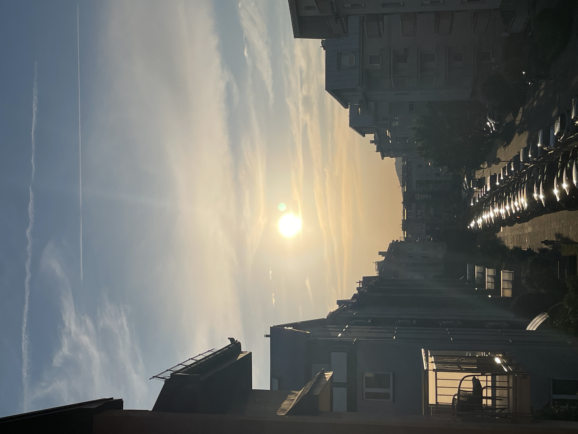 The image depicts an urban scene at sunset. The sun is low in the sky, casting a warm, golden light that bathes the buildings and sky in a soft glow. The sky is a gradient of blue and yellow, with wispy clouds and contrails from an airplane visible. In the foreground, the rooftops of several buildings are visible, with some showing architectural details like angled roofs and skylights. In the middle ground, a line of parked cars is illuminated by the setting sun, their reflections shimmering on the wet asphalt. The buildings in the background are silhouetted against the bright sky, with their windows reflecting the light. A lone bird is perched on the edge of one of the rooftops. The scene conveys a sense of calm and tranquility, with the warm light and peaceful atmosphere of a day drawing to a close. There is no visible text in the image.
