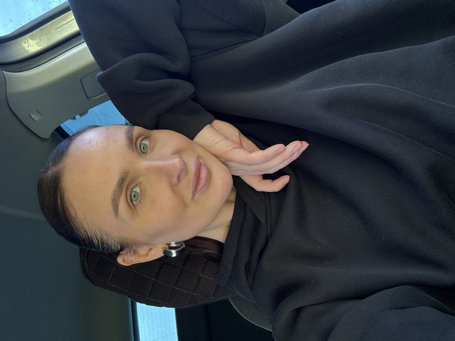 A close-up portrait shows a woman seated in what appears to be the front passenger seat of a vehicle. The woman has light skin, light green eyes, and dark hair styled neatly back from her face. She wears a black hooded sweatshirt and silver-toned hoop earrings. Her left hand is positioned with the back of her fingers resting against her right cheek/jawline. She is looking directly towards the camera with a subtle smile. Behind her, a black quilted headrest is visible. The vehicle's interior trim near the roofline appears light-colored. Through a rear window, a blurred, light blue background is discernible, suggesting an outdoor environment. The specific location within Chaikovsky, Russia, cannot be determined from the visual evidence within the image.