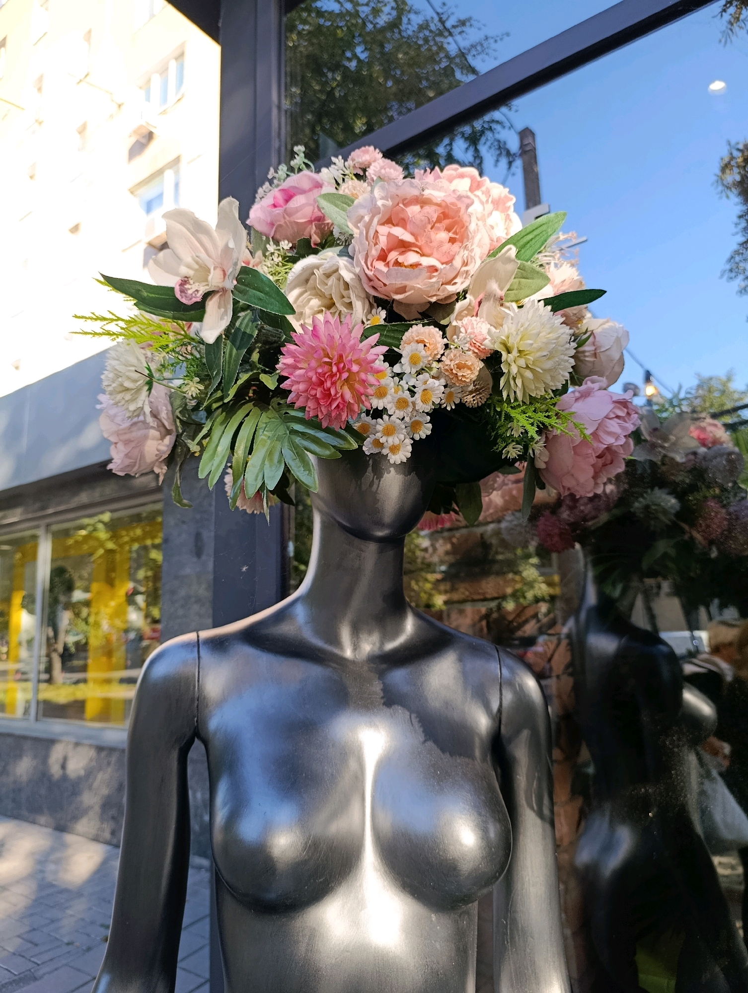 A black mannequin torso, displaying realistic human breast forms, stands outdoors on a paved walkway in front of a building with a glass facade. The mannequin is adorned with a large, elaborate floral arrangement on its head, featuring a variety of pink and white flowers, including peonies and daisies, with green foliage. The building's glass reflects the sky, trees, and other buildings, suggesting an urban setting. The sun casts shadows on the walkway, indicating it is daytime. The specific location of this scene is the display window of a flower shop on Khreshchatyk Street in Cherkasy, Ukraine, as evidenced by the building's architecture and storefront visible in the reflection. No individuals are present in the immediate vicinity of the mannequin.