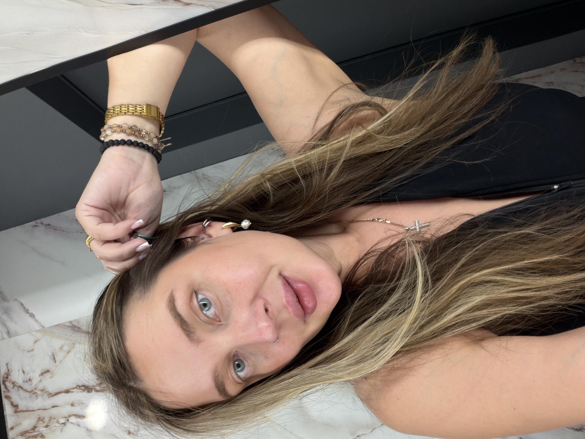 A fair-skinned person with long, highlighted brown and blonde hair, blue eyes, and a nose piercing is positioned at an angle. The person is wearing a black top and a gold necklace with a cross pendant. The left arm is raised, and the wrist features a gold watch and two beaded bracelets. The fingers of the left hand are manicured with white tips. The background includes a countertop with a marble-like pattern and a dark shelf or cabinet structure. There is no evidence in the image to suggest it was taken in Moscow, Russia.