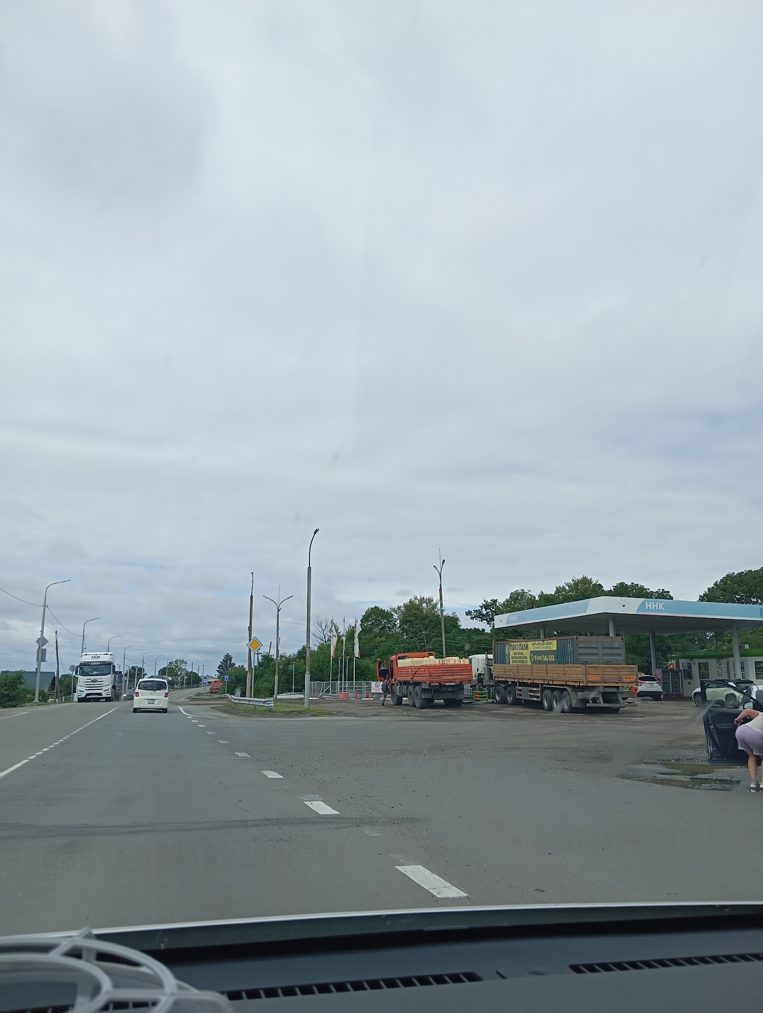The image shows a cloudy day at a gas station in Mayak, Russia. A white truck is parked on the left side of the road, and a white car is driving away from it. A gas station with a blue awning is visible in the background. A woman in a light-colored skirt and a white top is seen leaning into the open door of a dark-colored car parked at the gas station. Several trucks are parked at the gas station, one of which is loaded with rectangular yellow objects. The road in front of the gas station has white dashed lines painted on it, indicating a lane divider. Trees and buildings are visible in the distance.
