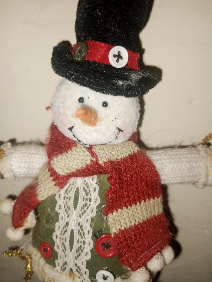 A charming snowman ornament is featured in the image. It has a black top hat with a red band and decorative buttons. Its face is a white, textured material with a carrot nose and stitched smile. It wears a red and tan striped scarf. The body of the snowman is adorned with an olive-green fabric and lace, embellished with buttons. The snowman is strung with gold tinsel.
