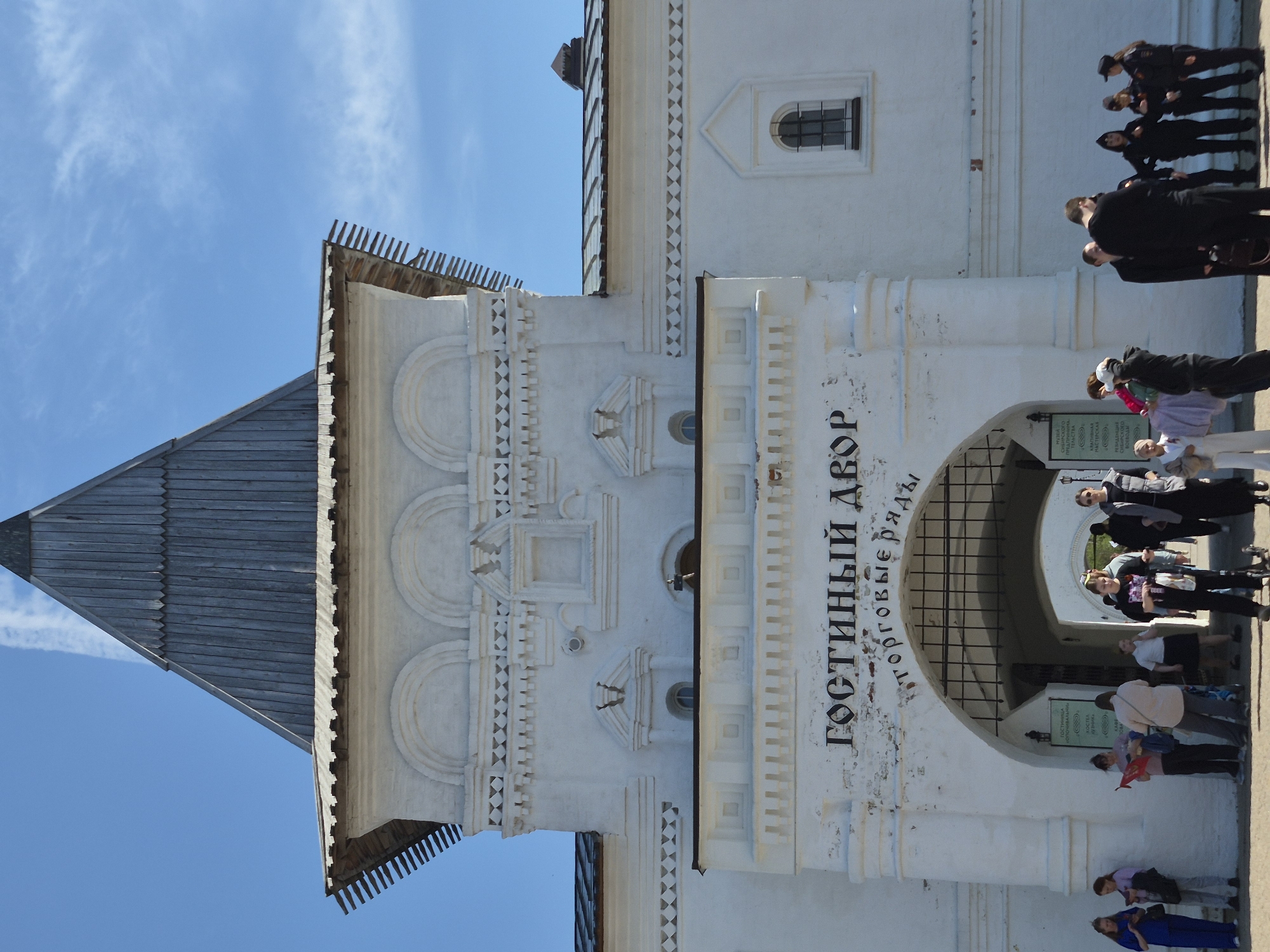 The image shows the Gostiny Dvor in Tobolsk, Russia, an architectural ensemble with a large white building, a wooden-roofed tower, and an arched entrance. A group of people is visible passing through the entrance, some walking and others standing. The sky is blue with some wispy clouds.