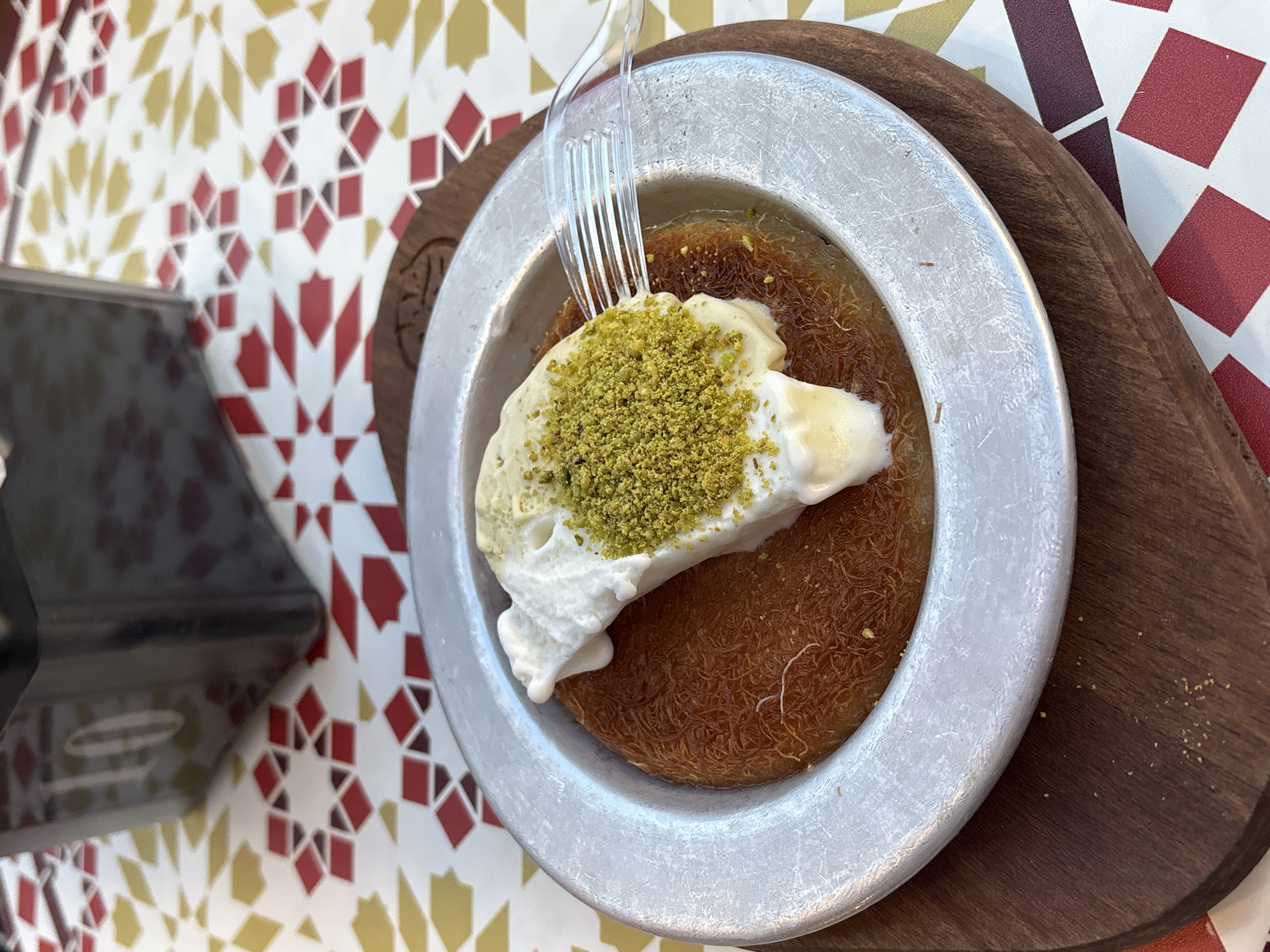 A close-up, overhead shot captures a plated dessert, likely Kunafa, resting on a wooden board. The dessert itself is a circular, golden-brown pastry topped with a dollop of white cream, generously sprinkled with chopped pistachios. A clear plastic fork is inserted into the cream, suggesting it has been eaten. The dessert sits on a metallic-grey plate with a slightly worn texture. The background features a colorful, patterned tablecloth with a repeating geometric design of red, gold, and white diamonds. To the left, a dark, square object, possibly a sugar dispenser or condiment holder, displays the number "10" in white. The lighting suggests an outdoor or brightly lit indoor setting. This dessert is presented at "Mishmish," a restaurant located at 7 King George Street in Tel-Aviv, Israel.
