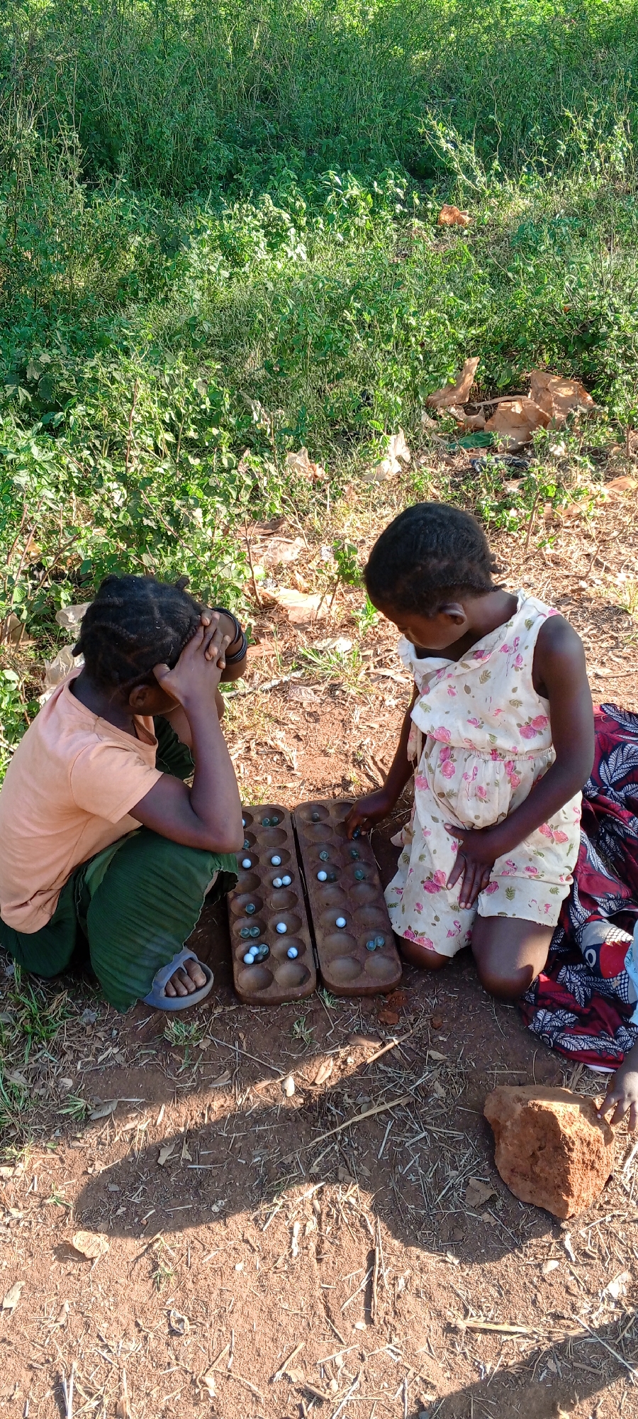 Two young girls are playing a game of mancala on the ground in an outdoor setting in Ngalula, Democratic Republic of the Congo. The girl on the left is wearing a peach-colored t-shirt and a dark green skirt. She has dark hair and is resting her head in her hands, seemingly deep in thought. She is wearing a pair of light-colored sandals. The girl on the right is wearing a light-colored floral dress and is kneeling on the ground, reaching out to move a piece in the game. Between them is a wooden mancala board with two rows of six holes each. Some of the holes contain white marbles, while others are empty. To the right of the kneeling girl is a red and black patterned cloth. The ground is dirt with patches of grass. Behind the girls is an area of dense green vegetation. A red rock is visible in the lower right corner of the image.