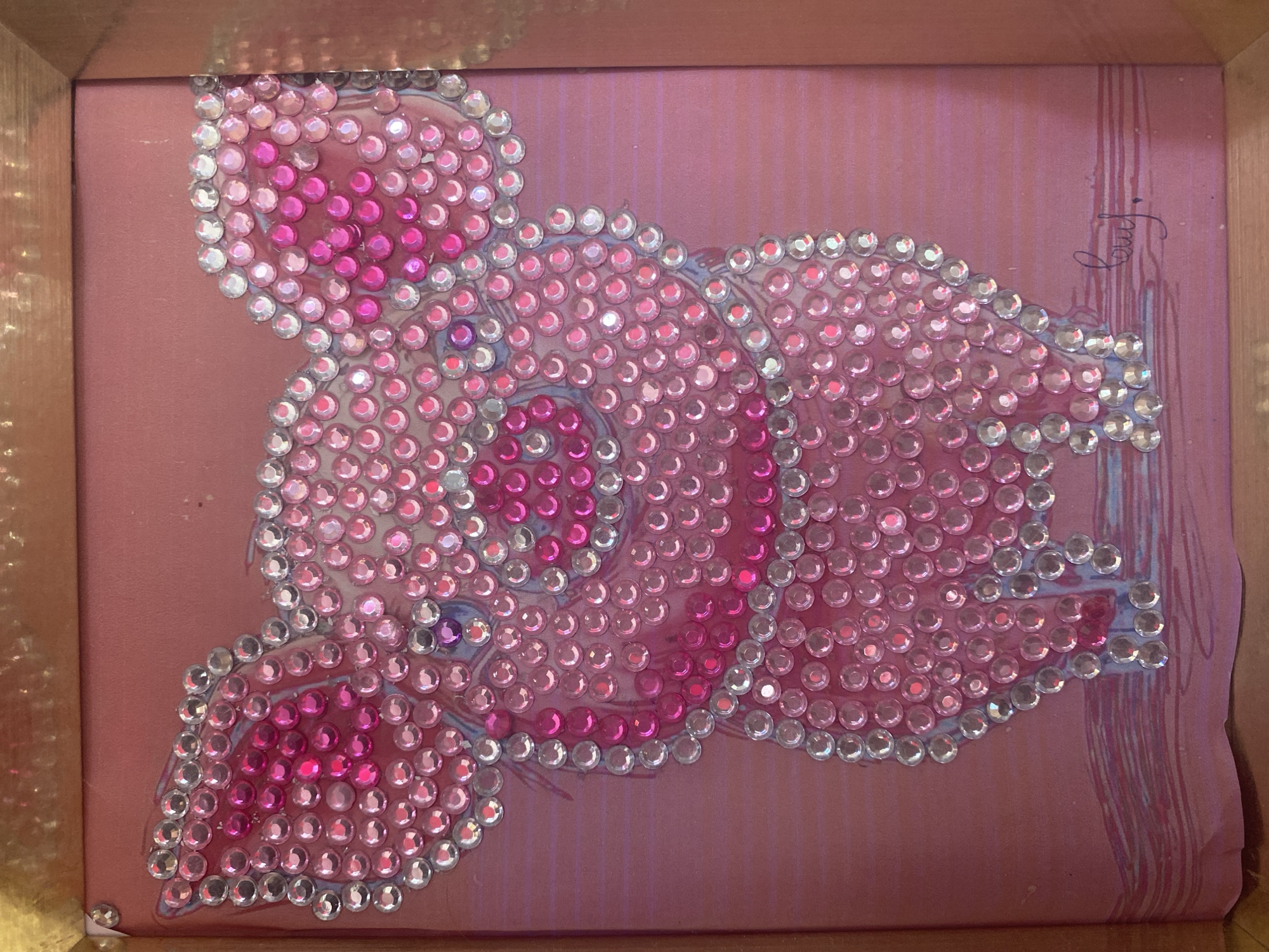 A framed craft art piece depicts a pink piglet adorned with numerous small, clear and pink rhinestones. The piglet is drawn on a pink background with some subtle horizontal lines. The art is contained within a light brown frame, which is slightly ajar in the top left corner. A faint blue signature is visible on the bottom right of the artwork. The image was taken indoors, likely in a home or studio setting. No specific location within Blagoevgrad, Bulgaria can be determined from the image.