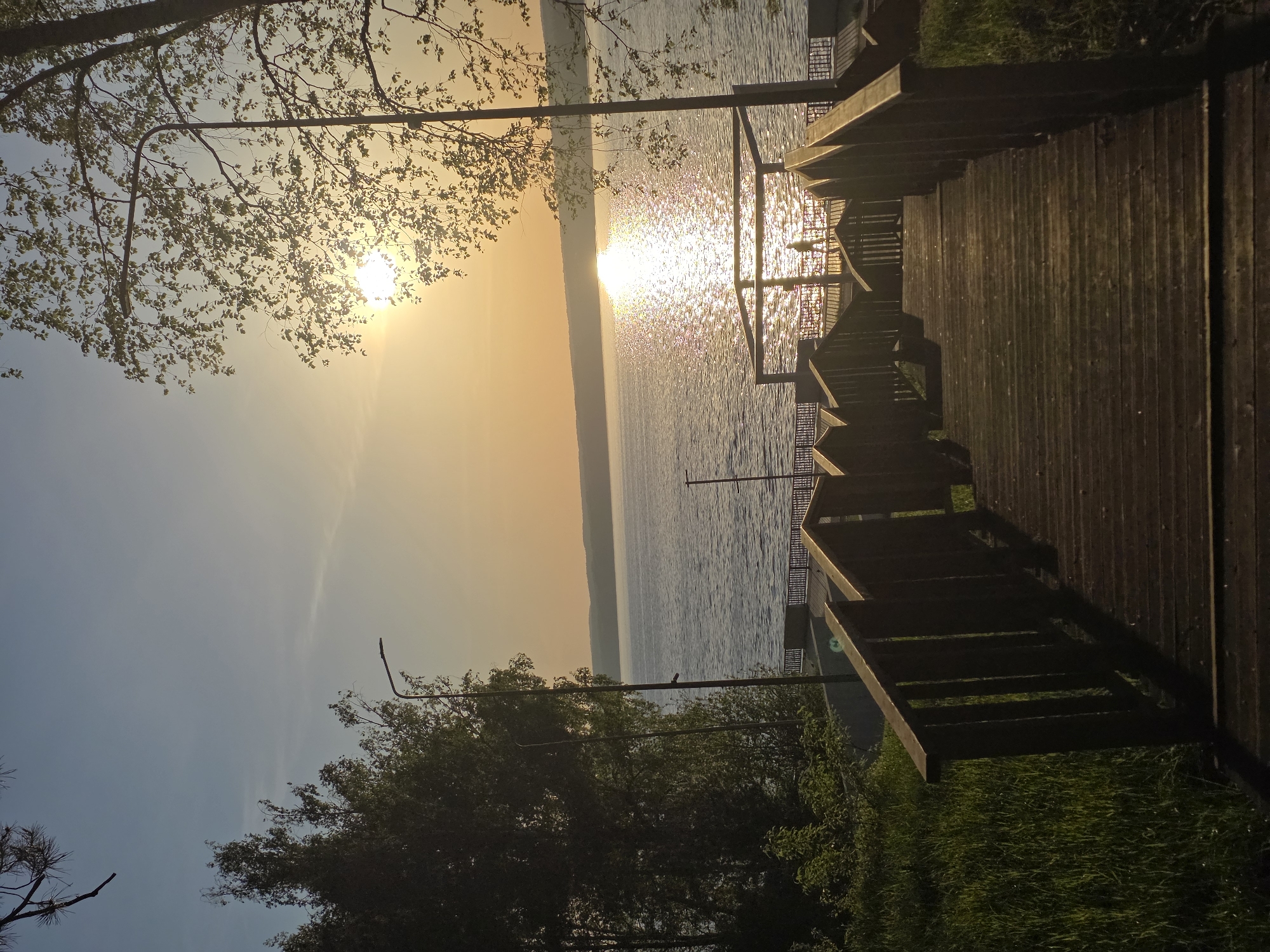 The image depicts a sunset view over a body of water, likely a lake or river, from a wooden walkway with railings and steps. The sun is low on the horizon, creating a strong glare and reflection on the water surface. Trees and foliage frame the view on both sides. A lamp post is visible near the upper left corner, partially obscured by tree branches. The sky transitions from a lighter blue in the lower left to a golden hue near the sun. The scene is probably in Chaikovsky, Russia.