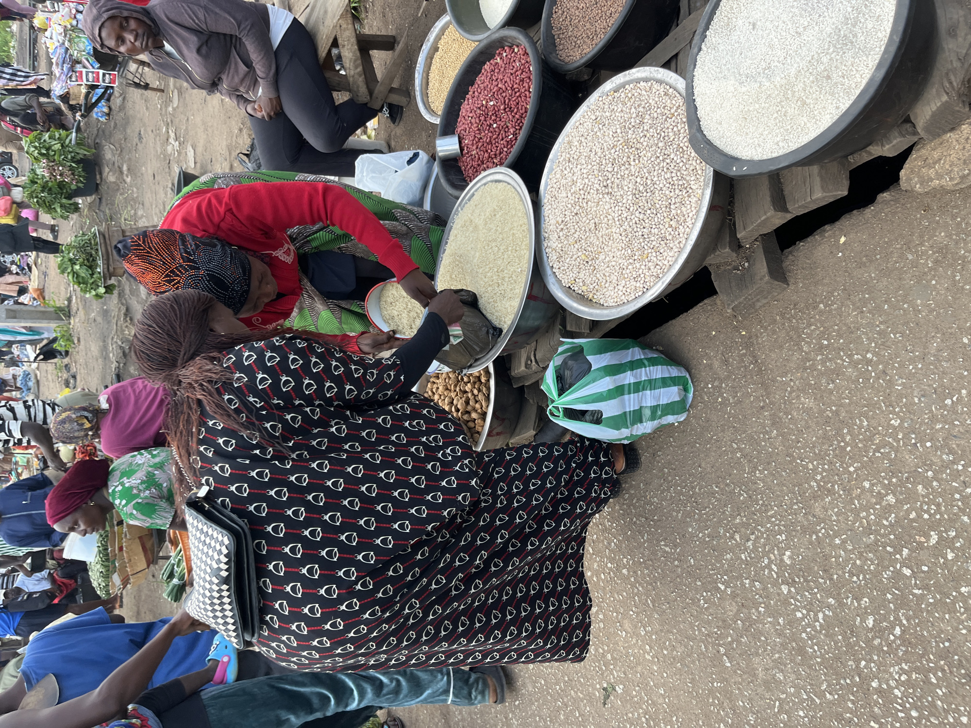 At a market in Bukuru, Nigeria, a group of women are engaged in commerce. One woman, wearing a black dress adorned with white and red patterns of horse bits, is seen from the back and side as she interacts with a vendor. The vendor, in a bright red top and a colorful headscarf, is scooping white grains, likely rice, from a large metal basin into a black plastic bag. Another woman, to the left, is wearing a green and white patterned dress and a red headscarf, her face turned away from the camera. A fourth woman, seated on a wooden bench in the background, wears a hooded brown jacket and dark pants, her gaze directed downwards. The market is bustling with activity, evidenced by the presence of various food items displayed in large basins. These include what appear to be peanuts, various types of beans, and grains such as rice and millet. The ground is paved with rough, uneven asphalt, suggesting a typical outdoor market setting. The overall atmosphere suggests a vibrant and active marketplace.