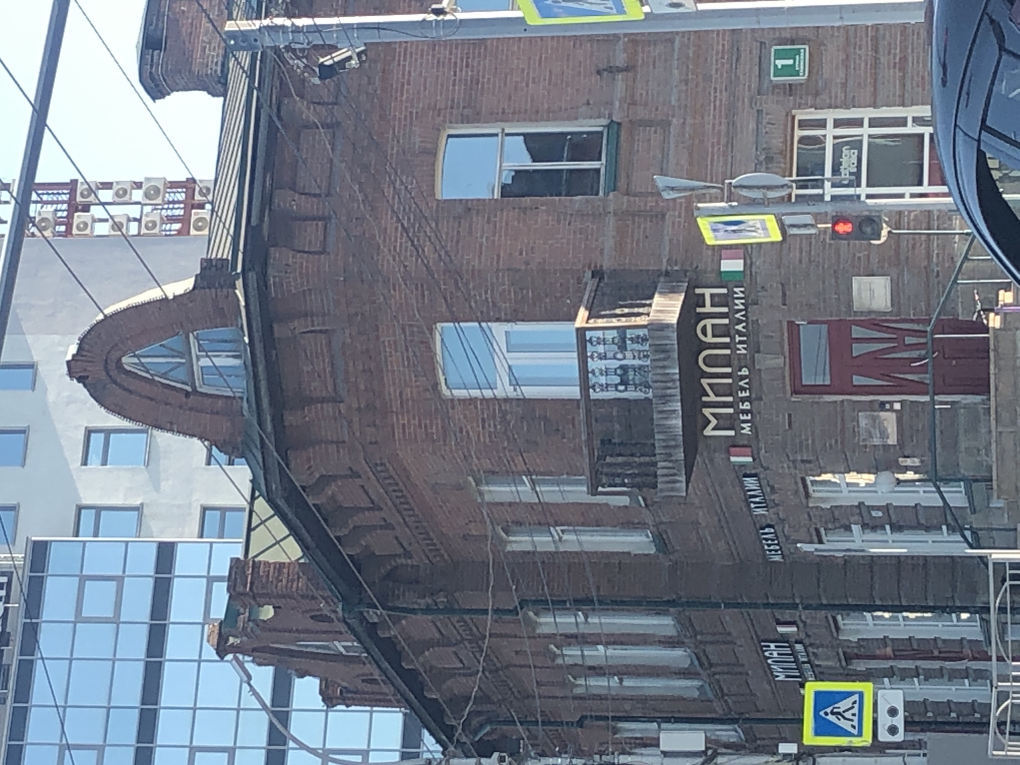 The image shows a brick building with arched windows and a small balcony. There are signs on the building that read "МИЛАН" and "МЕБЕЛЬ ИТАЛИИ", indicating a Milanese furniture store. A pedestrian crossing sign and a traffic light are visible on the street in front of the building. In the background, a modern glass building and several air conditioning units on a red structure can be seen. The scene appears to be a street in an urban environment, with a car visible in the foreground.