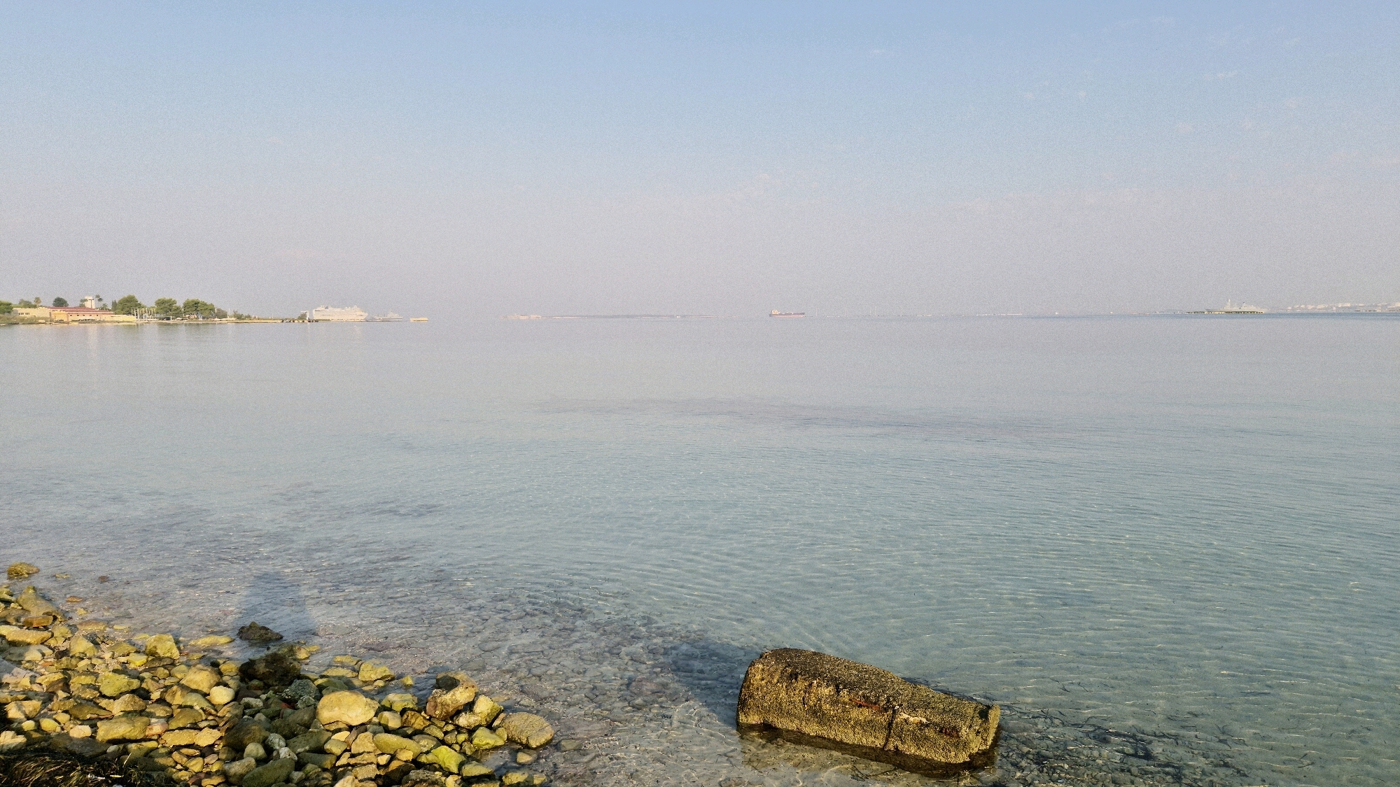 The image depicts a serene coastal scene in Taranto, Italy, likely from the Mar Piccolo. In the foreground, a rocky shoreline is visible, with various sized stones, some appearing moss-covered, leading to the clear, shallow water. A large, rectangular object, possibly a concrete block, rests partially submerged in the water near the shore. The water itself is calm and transparent, revealing the seabed.
In the middle ground, the vast expanse of the sea stretches out, with a hazy horizon. Several vessels are discernible in the distance, including what appears to be a large cruise ship or ferry anchored near a landmass on the left. Further out, other ships, including a cargo ship, are scattered across the water.
The background features a faint coastline with buildings and trees, hinting at the urban landscape of Taranto. The sky is a clear, pale blue, suggesting a bright, possibly morning or late afternoon, lighting condition. The overall atmosphere is peaceful and tranquil, capturing the essence of a coastal view in Taranto.