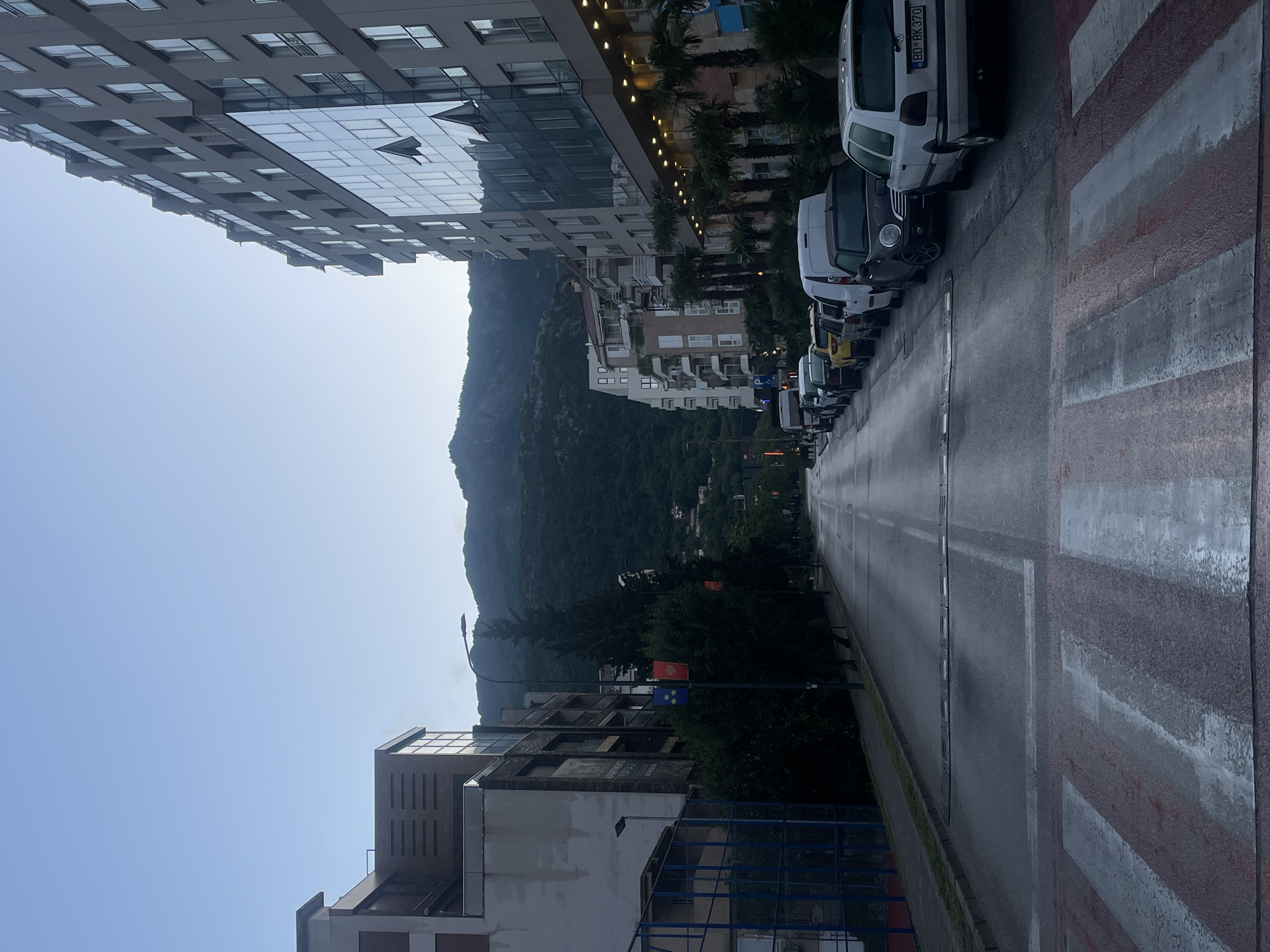 The image depicts a street scene in an urban environment. On one side of the street, a modern building with reflective glass windows rises, and on the opposite side, there's a mix of older buildings. Parked vehicles line the street. In the background, there's a lush green mountain, suggesting the city is located near a natural landscape. A pedestrian crossing is visible in the foreground. National flags and lampposts are visible on the side of the street. The sky is bright, suggesting daytime. The location appears to be in Montenegro, given the vehicles licence plate and flags.