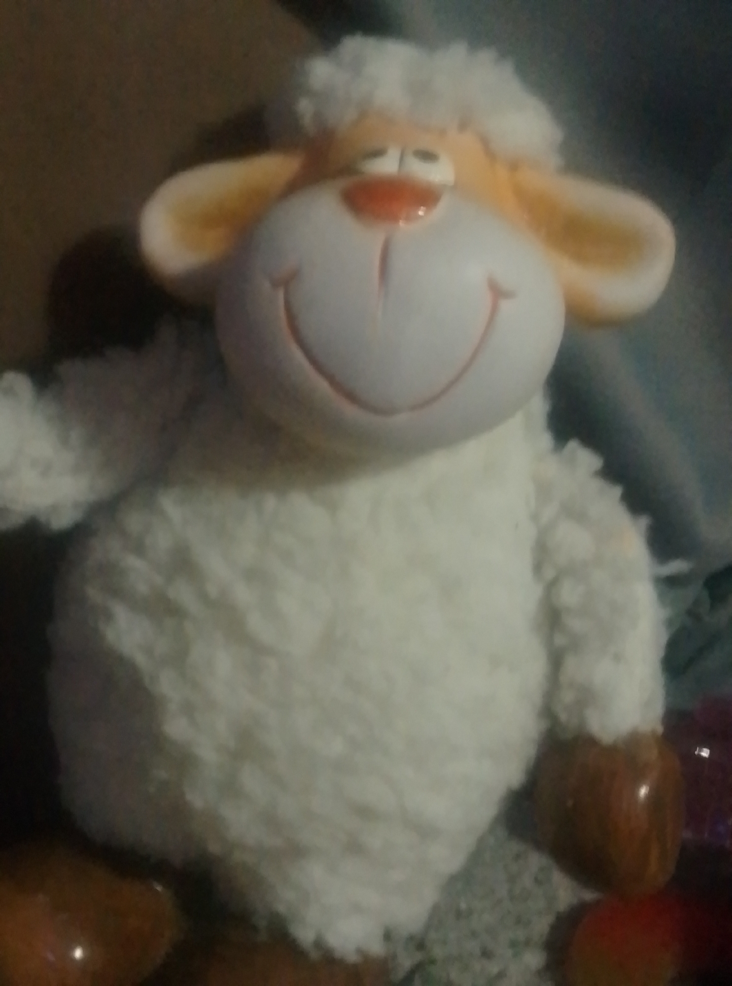 A plush toy sheep is displayed against a neutral background. The sheep is white with a fluffy texture, featuring a cream-colored face with orange accents on its nose and ears. It has closed eyes and a smiling mouth. The toy's feet are a brown, glossy material. The lighting is dim, creating shadows and a slightly blurry image. The scene is indoors. The image does not depict any specific location in the city of Wedel, Germany.