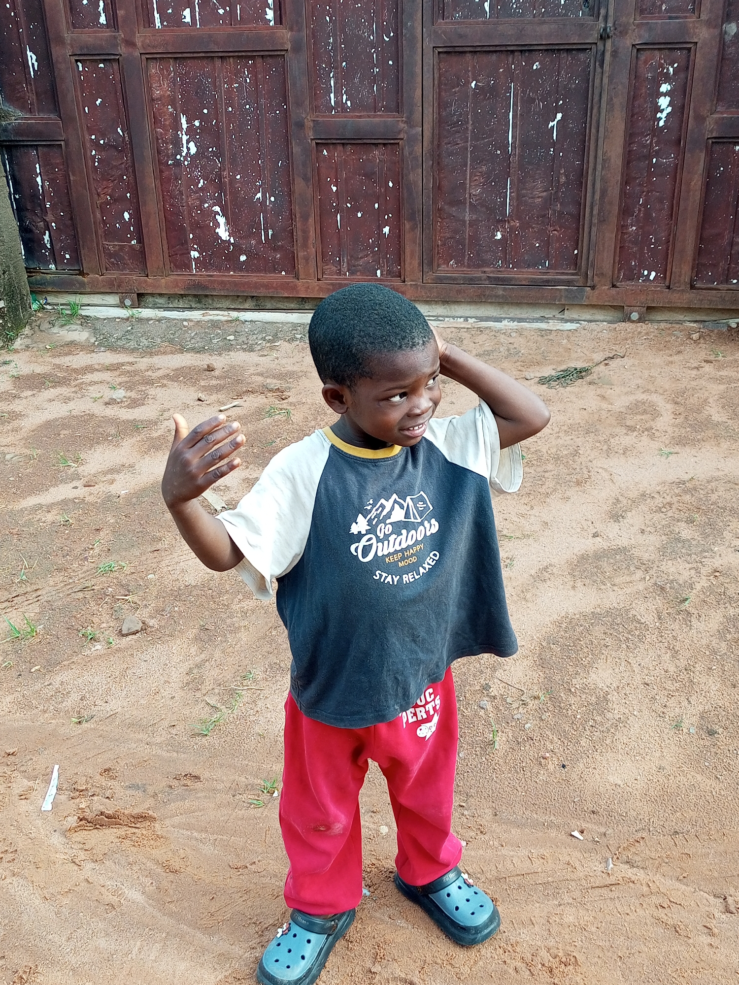 A young boy with dark skin and short, dark hair stands in an outdoor setting, looking to his right with a slight smile. He wears a dark t-shirt with "Go Outdoors Keep Happy Mood Stay Relaxed" printed on it, and red pants. His right hand is raised to his head. He is wearing dark blue Croc-style shoes. The ground is covered in sand with scattered green grass and small rocks. Behind him is a large, dark brown, weathered wooden gate with vertical planks and white splotches. The scene appears to be in Tempure, Nigeria.