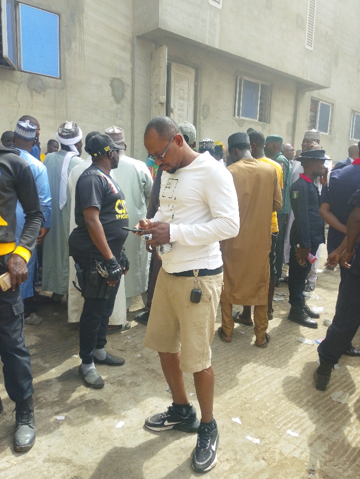 An outdoor scene in Fagge C, Nigeria, depicts a gathering of approximately 15-20 adult males in front of an unfinished or unpainted concrete building. The building features multiple rectangular windows with blue panes, some displaying security bars, and two plain white doors. The ground is a light-colored, possibly concrete or dirt, surface littered with small pieces of white paper.
In the foreground, slightly right of center, a light-skinned male with a short beard and glasses is seen. He wears a white long-sleeved t-shirt with a faint graphic, khaki shorts, and black Nike sneakers, and is actively engaged with a black smartphone held in both hands, looking down at the screen. A black accessory is clipped to his right belt loop.
To the immediate left of this central figure, a dark-skinned male wears a black t-shirt with yellow "SPECIALIST" lettering, black tactical pants, black tactical gloves, and a black cap. A black tactical holster rig containing what appears to be a firearm is visible on his right hip. He faces slightly right.
The background and mid-ground are populated by a crowd of men, many wearing traditional Nigerian attire, including long robes (brown, light blue, green) and various white and patterned head coverings. Several individuals are in dark-colored uniforms, including one on the far right with a peak cap and black boots, indicative of security or police personnel. A person in a yellow top is also visible in the crowd. The individuals are standing in a general gathering; no specific interactions are overtly discernible beyond their presence in proximity. Natural daylight illuminates the scene.