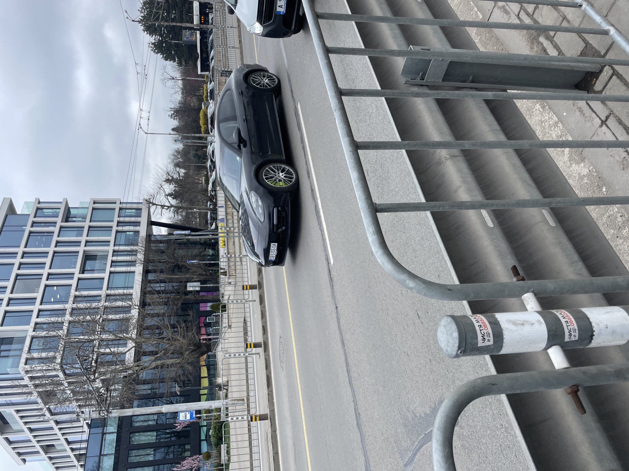 The image depicts a street scene in Sofia, Bulgaria, featuring a modern building with a grid-like facade on the left side of the frame. A black Porsche is driving on the road, and a white van is in front of it. Metal pedestrian barriers line the road. The sky is overcast. The license plate of the Porsche appears to read "CB 5155 OA".