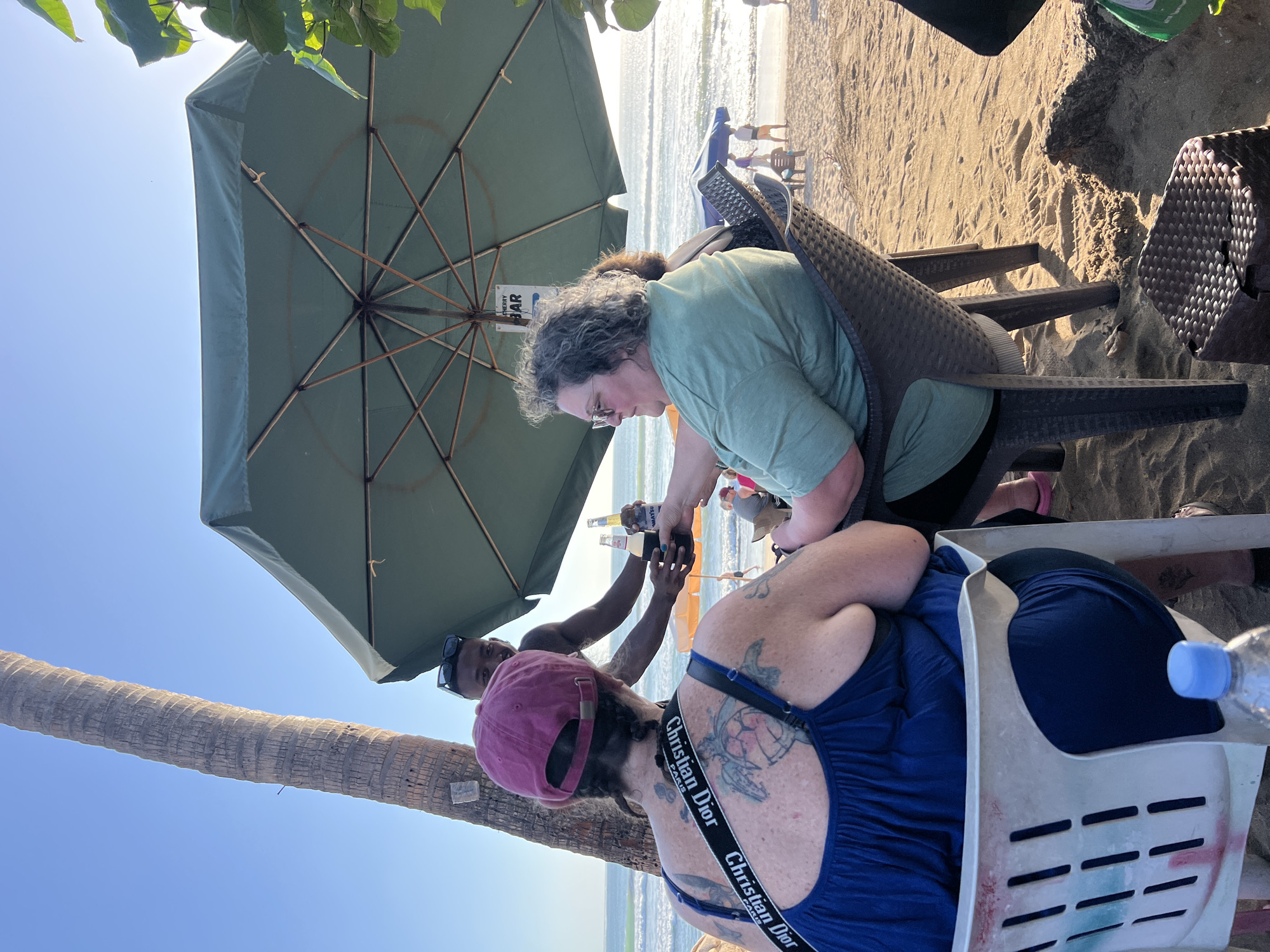 Two women and a server are seated at tables on a beach in Kuta, Indonesia, under a large green umbrella. The woman on the left is wearing a blue tank top and a maroon baseball cap, with tattoos on her back and arm. The woman on the right is wearing a light green t-shirt and glasses. The server, a man with sunglasses, is handing the women two bottles of beer. The ocean is visible in the background, with waves and several people walking on the beach. Palm trees and lush green foliage frame the scene. The sand is golden brown, and there are footprints and debris scattered across it.