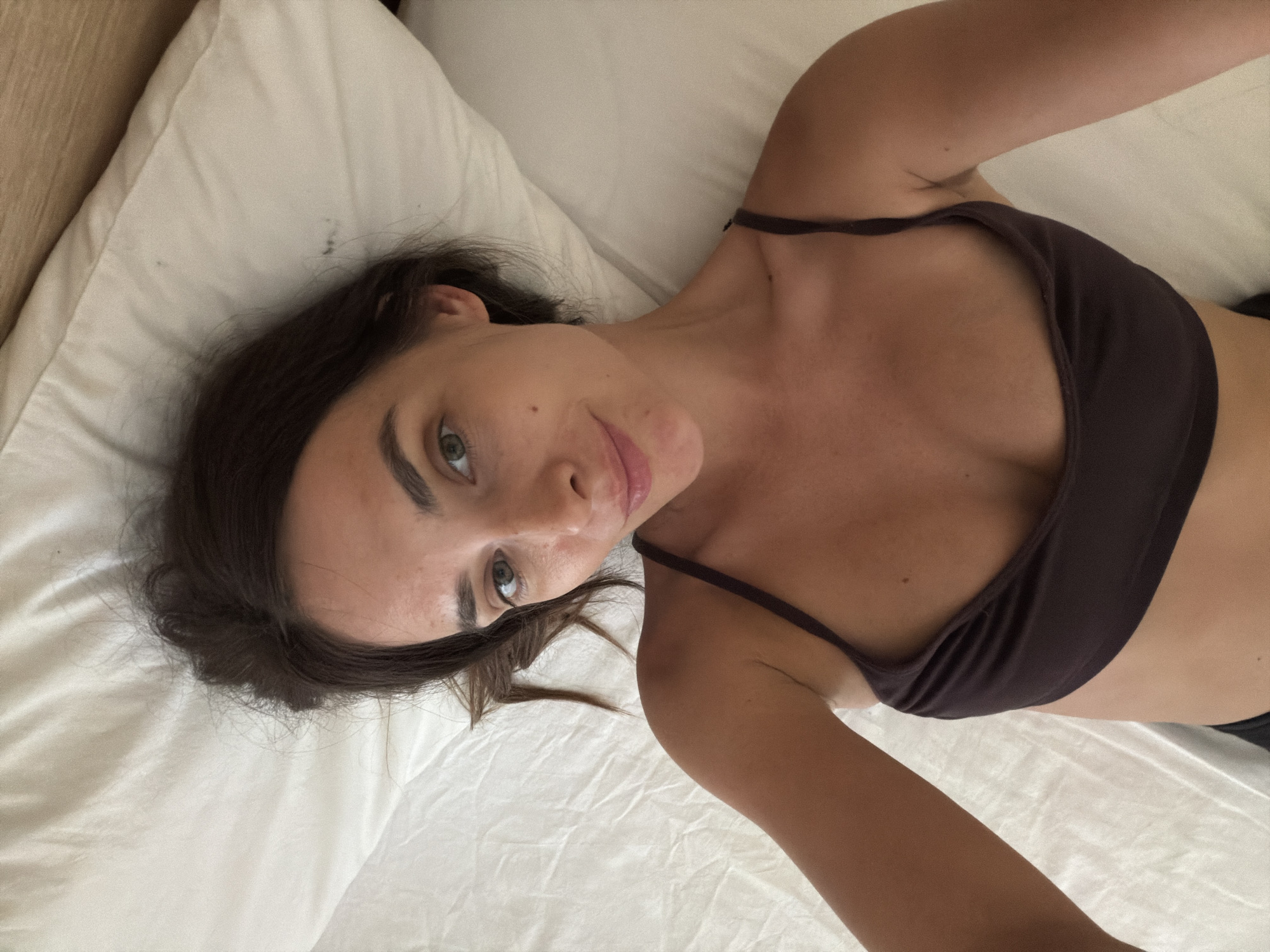 A woman is lying on a white bed with her head on a white pillow. She is wearing a dark brown camisole. Her hair is dark brown and falls around her face and shoulders. Her eyes are green and she is looking at the camera with a slight smile. The room is dimly lit, suggesting it might be early morning or late evening. The sheets are wrinkled, indicating the bed has been slept in. The overall mood is relaxed and intimate, a private moment captured in what appears to be a bedroom. The location is Fethiye, Turkey.