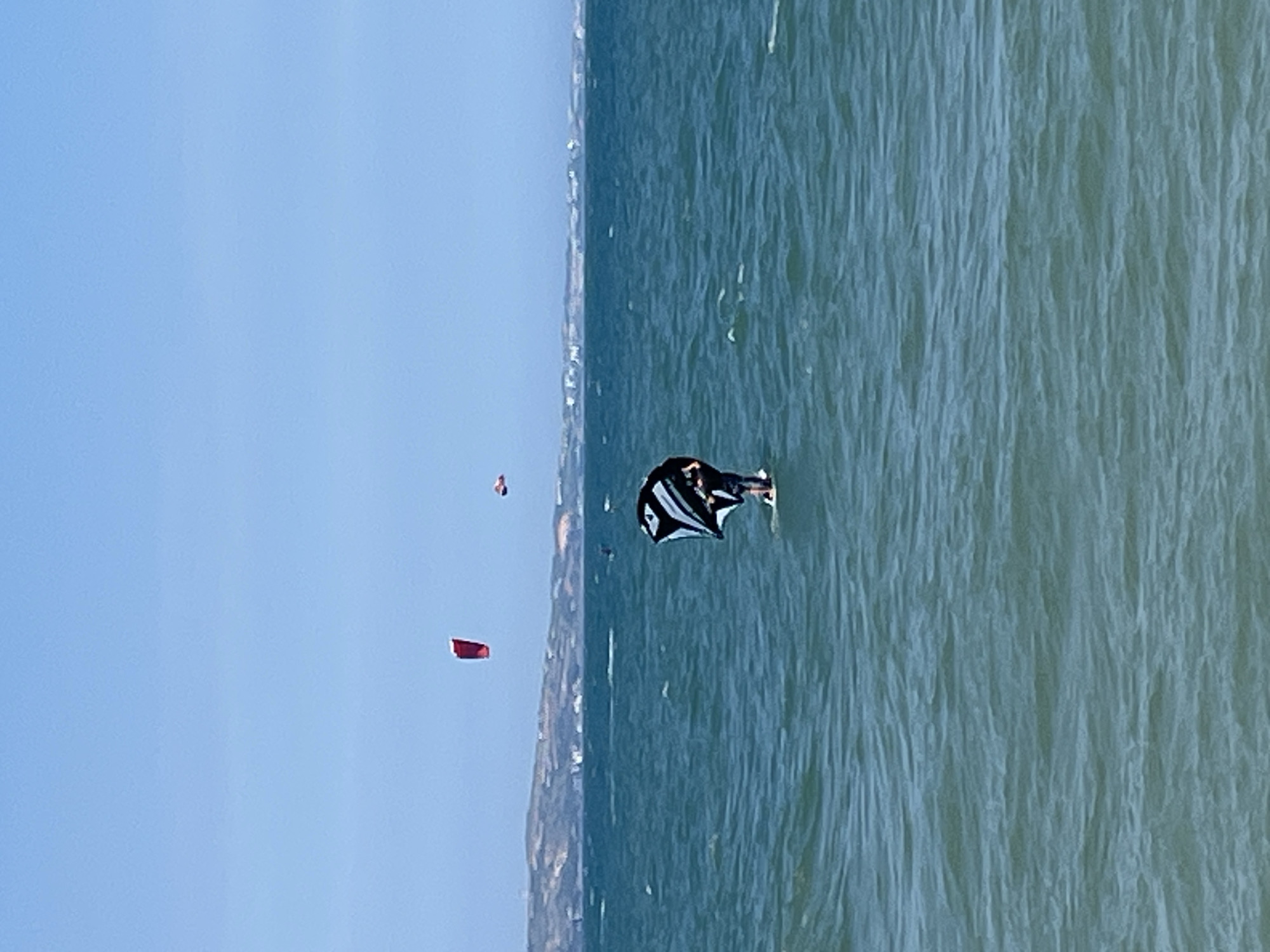 In the image, a person is kiteboarding on a body of water. The kiteboarder is wearing a dark shirt and shorts and is standing on a board. They are holding a black and white kite. The water is choppy and a deep blue. In the background, there is a shoreline with hills and buildings. The sky is clear and blue. There are other kites in the sky, one red and one orange. The location is Phu Thuy Ward, Vietnam.