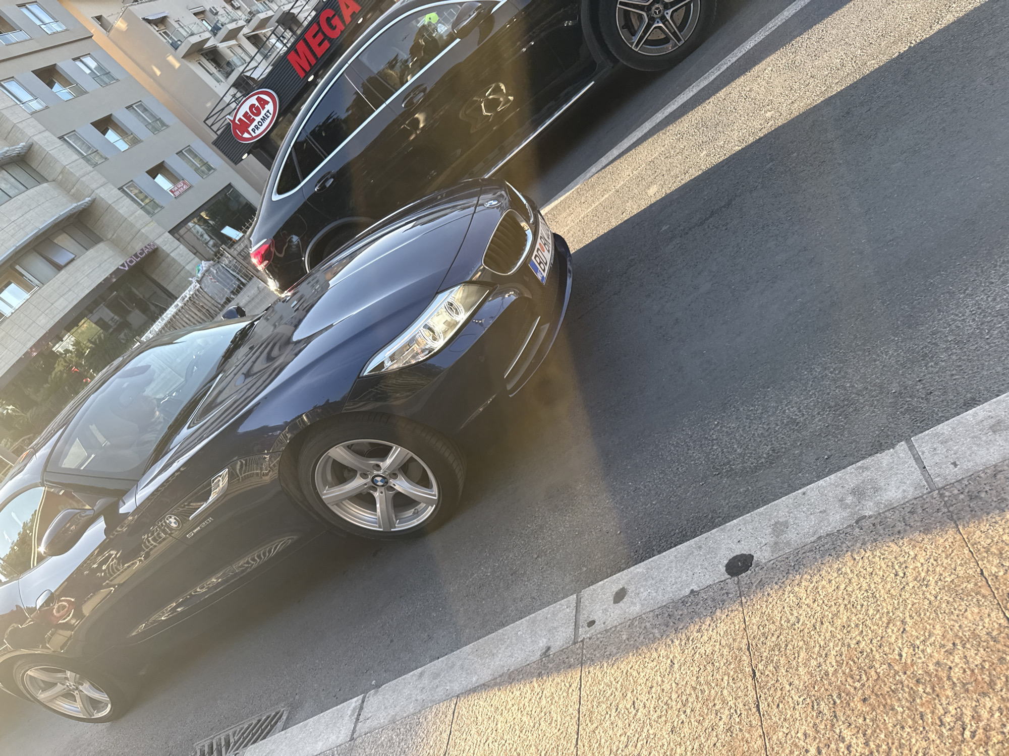 A photograph shows a dark blue BMW Z4 Roadster parked on a street in Budva, Montenegro. The car has silver rims and a license plate with "BD" visible. A black Mercedes SUV is parked next to it, partially obscuring the view of the building behind. The building has a "MEGA PROMET" sign. The photograph has a warm, sunny light, casting shadows on the street and sidewalk. The street is paved and the sidewalk is made of rectangular stones. The photograph appears to be taken at street level.