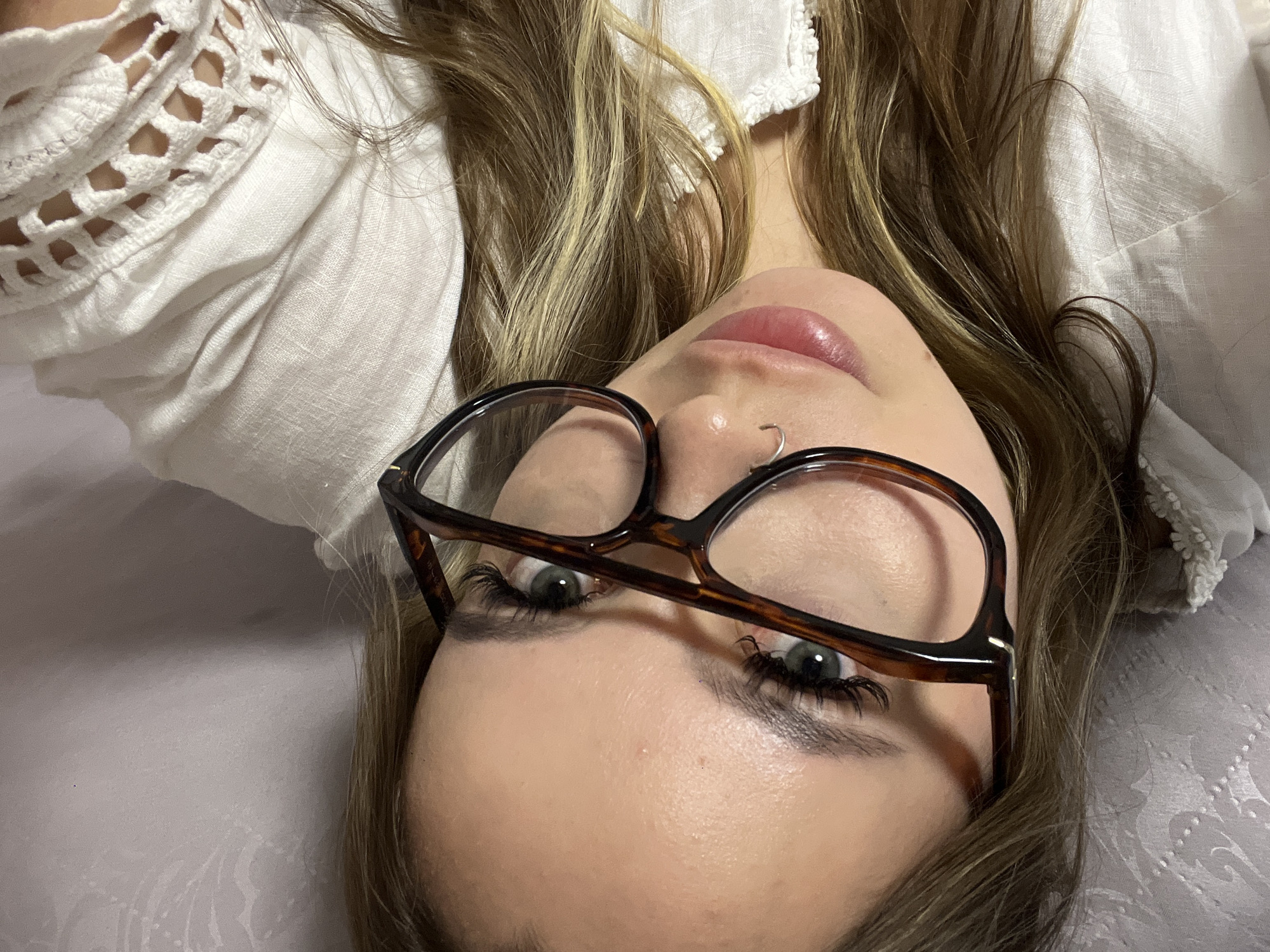 A young woman with blonde highlighted hair is lying on a textured light-colored surface, possibly a bed or pillow. She is wearing glasses with tortoise-shell frames and a white, linen-textured shirt with lace detailing. She has a small silver nose ring and appears to be looking directly at the camera, although the image is rotated. There is no indication of the location in the city of Krasnodar, Russia. The environment is nondescript.
