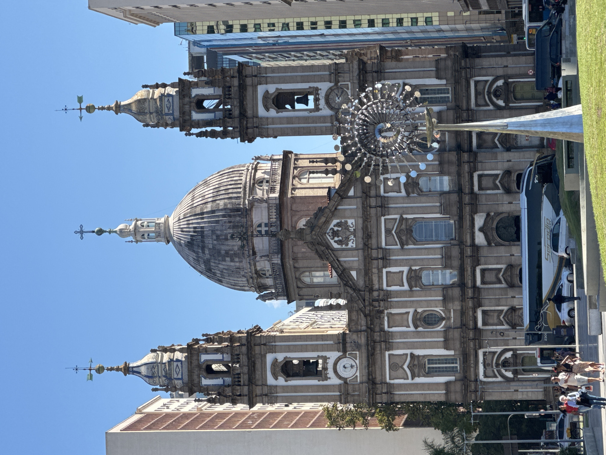 The image shows the Candelária Church in Rio de Janeiro, Brazil. It is a large, neoclassical church with a prominent dome and two bell towers. The church is flanked by modern buildings, and in front of it is a busy street with cars and pedestrians. A bus and several cars are visible on the road. There is also a modern sculpture in the foreground. The sky is clear and blue. Several people are walking on a sidewalk in front of the church, some carrying bags.