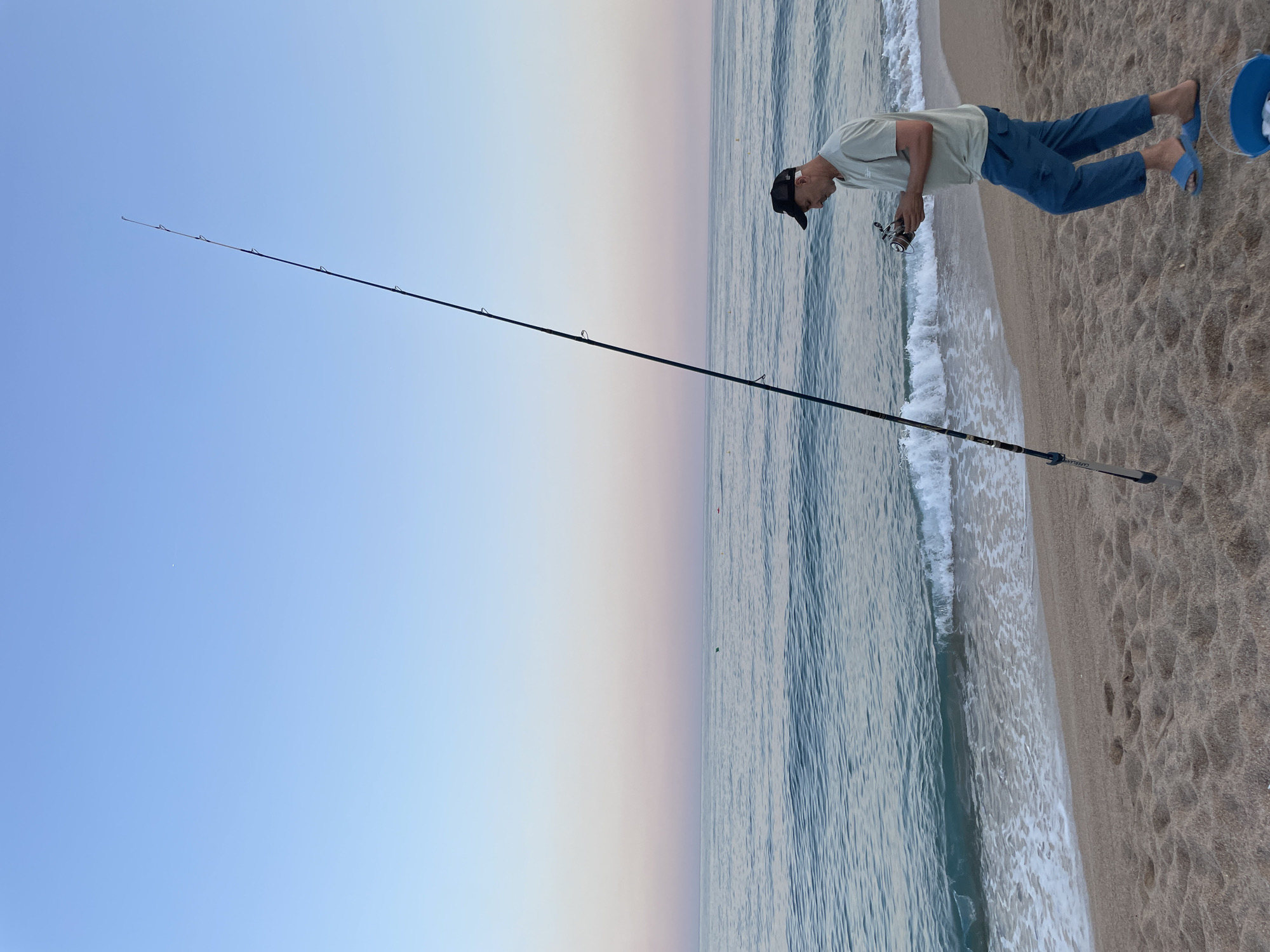 A man stands on a sandy beach in Canet de Mar, Spain, near the waterline, holding a fishing reel. He is wearing a light green t-shirt, blue pants, a black baseball cap, and blue flip-flops. A long fishing rod is planted in the sand beside him, angling upwards towards the clear sky. The ocean is calm with gentle waves lapping at the shore. The sky displays a gradient of soft blue and pink hues, indicating it is either sunrise or sunset. A blue bucket with some white objects inside sits on the sand to the man's right. The overall scene captures a serene moment of recreational fishing by the Mediterranean Sea.