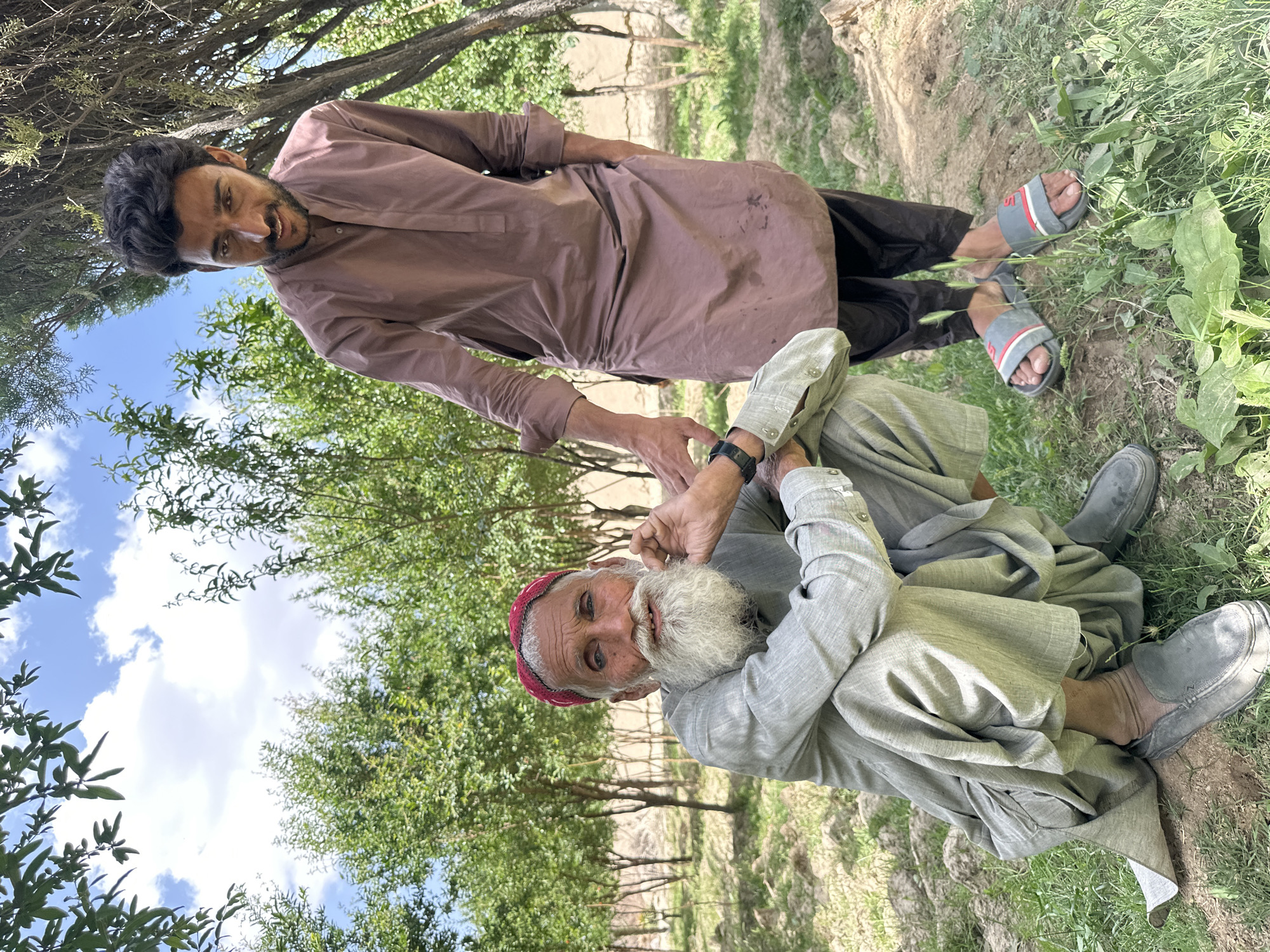 Two men are in an outdoor setting. An older man with a white beard and a red hat is seated in a grey traditional Pakistani dress. A younger man stands behind the seated man wearing a brown tunic and black pants. The younger man is holding the hands of the seated man. The background includes trees, grass and a blue and white sky. The location is Quetta City Tehsil, Pakistan.