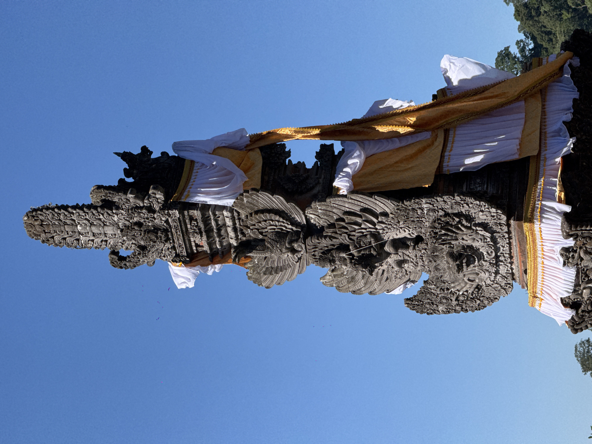 A stone statue with intricate carvings of mythical creatures stands against a clear blue sky in Baturiti, Indonesia. The statue is adorned with white and yellow fabric draped around its structure. The fabric is pleated at the bottom, creating a decorative effect. The statue appears to be part of a larger temple or shrine complex, as suggested by its ornate design and the surrounding foliage visible in the lower right corner. The overall scene conveys a sense of cultural and religious significance.