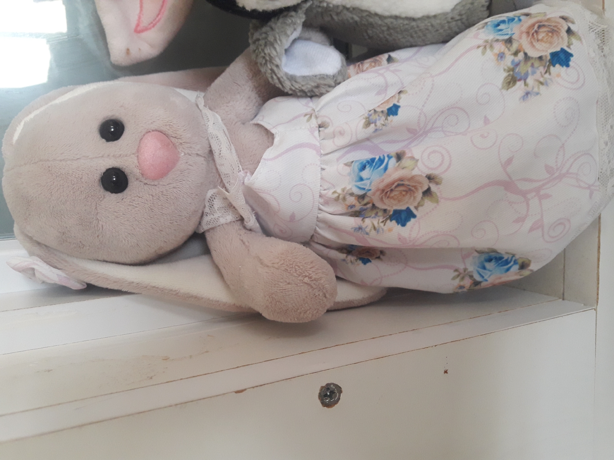 A close-up, low-angle shot shows a stuffed rabbit in a white dress with a floral pattern and lace trim, standing next to other stuffed animals. The rabbit is light gray with long ears, black eyes, and a pink nose. The dress has a light pink floral pattern with blue and beige roses. Behind the rabbit, a portion of another stuffed animal, possibly a penguin or another rabbit, is visible. The background consists of a white wooden surface with visible seams and some minor imperfections. There is a screw visible on the left side of the white surface. The lighting is soft, casting subtle shadows and highlighting the textures of the stuffed animals and the wooden surface. The scene is devoid of any human presence or activity.