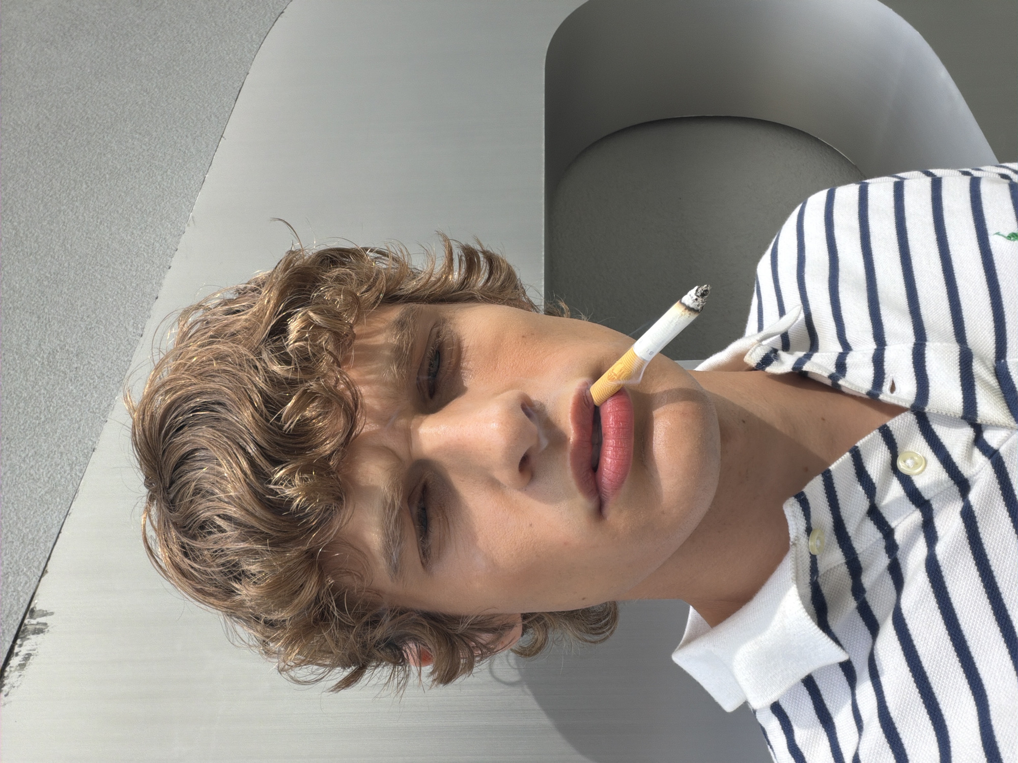 A Dutch-angled close-up portrait captures a young individual, likely male, with light skin and curly, light brown hair, set against an abstract, minimalist background. The subject's head is tilted significantly to the left, with their eyes partially closed or looking downwards, conveying a relaxed or contemplative expression. A partially lit cigarette, with a visible filter and ash, is held between their slightly parted, pink lips.
The individual wears a white polo shirt adorned with distinct dark blue vertical stripes. A small, embroidered green logo, resembling a polo player, is visible on the left side of the shirt.
The background is composed of various shades of grey and white, featuring smooth, curved architectural elements that suggest modern design, such as an archway or a curved wall. One section appears to be a textured, lighter grey surface. Bright, natural light illuminates the scene, casting strong highlights on the subject's hair and skin, and creating subtle shadows within the geometric background, indicative of a clear, sunny day. The setting suggests a contemporary urban or architectural environment, consistent with a location like Milan, Italy. No explicit text is visible in the image.