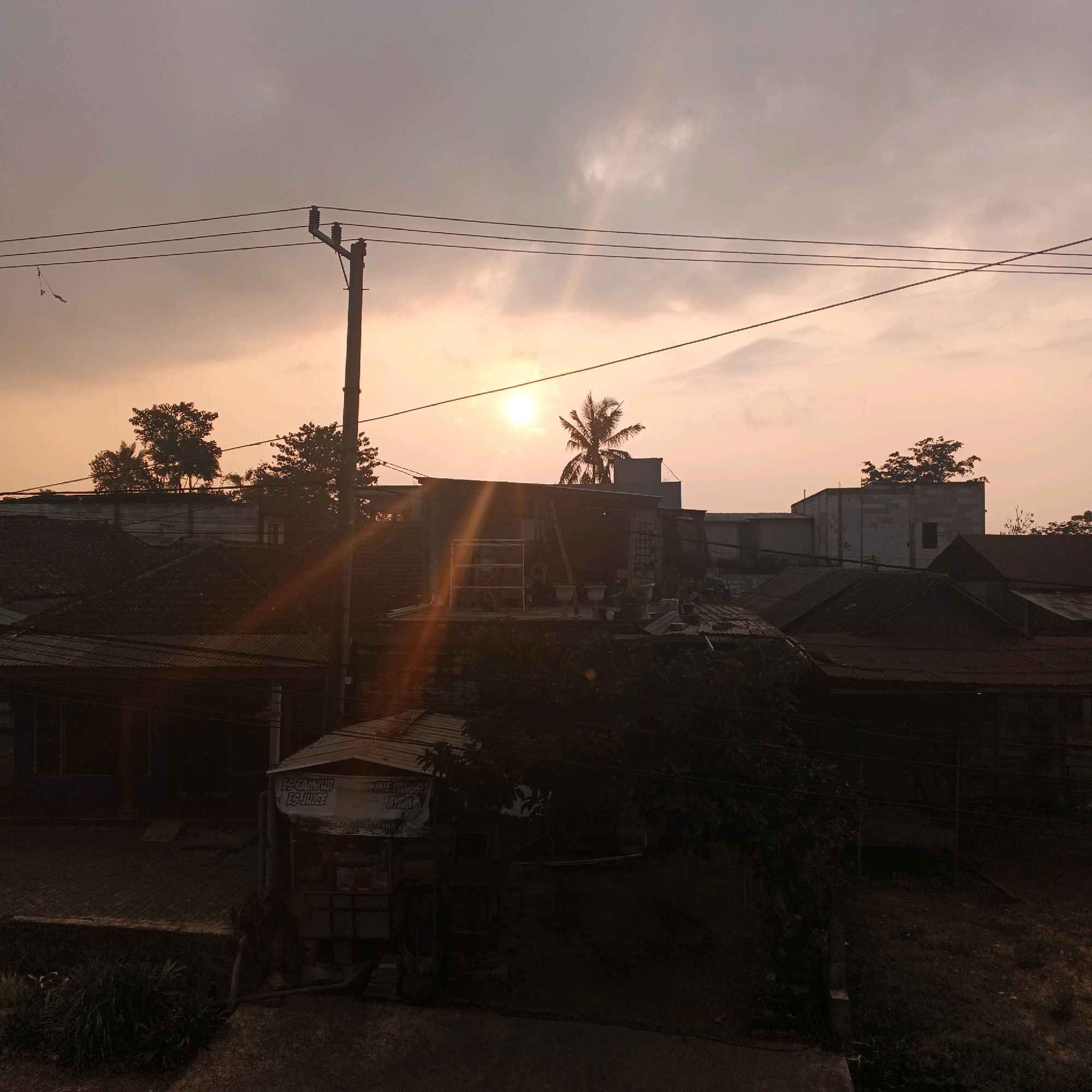 The image depicts an outdoor scene at sunset in Bocek, Indonesia. In the foreground, there are several dark, silhouetted structures, likely residential homes with tiled roofs, and a small roadside stall with a banner advertising "ES CAMPUR ES JUICE" and "MI TING AYAM". A dirt or gravel path is visible in front of the stall. Overhead, a utility pole with multiple wires stretches across the frame. The middle ground features more residential buildings and trees, including a palm tree. The background is dominated by a hazy, orange-hued sky, characteristic of a sunset, with the sun partially obscured by clouds and a lens flare creating streaks of light. The overall impression is a somewhat overcast sunset over a neighborhood with visible power lines and a small commercial establishment. No individuals are clearly visible in the image.