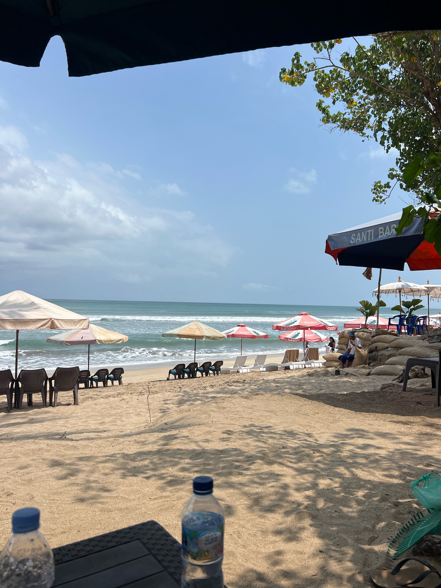 This image depicts a beach scene in Kuta, Indonesia, likely near a bar or cafe given the "Santi Bar" signage on an umbrella. In the foreground, two plastic water bottles sit on a dark, textured table, casting shadows on the sandy ground. Beyond the table, the sandy beach stretches towards the ocean, lined with rows of empty lounge chairs and umbrellas. The ocean has moderate waves and the sky is partly cloudy. In the middle ground, several people are visible, some seated and others standing near beach umbrellas. A man is sitting on a pile of sandbags. In the background, lush green foliage from trees hangs over the scene, providing shade. The overall atmosphere is one of a relaxed beach environment, possibly during a warm, humid day.