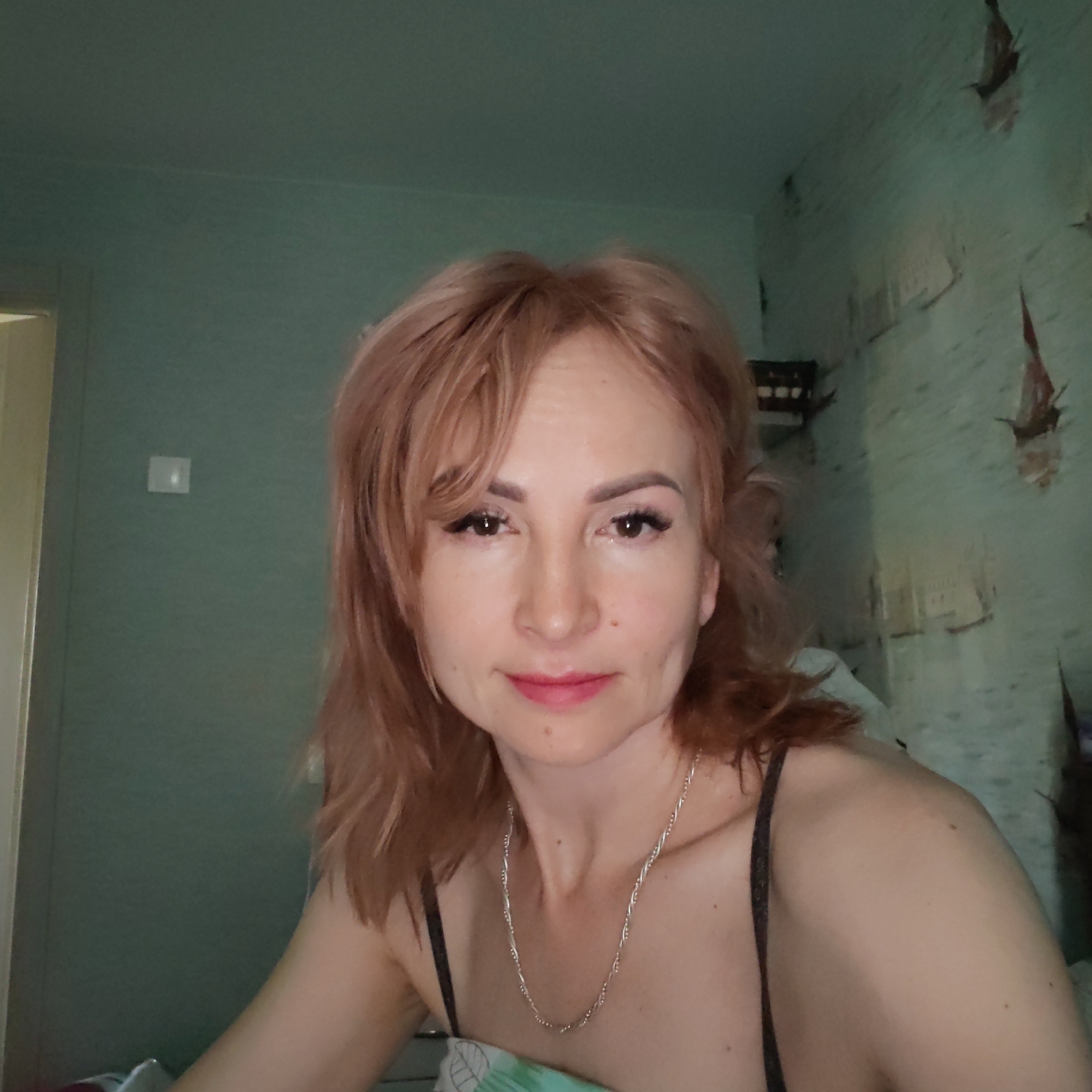 A middle-aged woman is in a room with green walls and a light switch on one wall. The wall behind her is light blue with ripped wallpaper. She has reddish-brown hair and is wearing a black tank top and a silver necklace. She is lying in a bed with a green and white pillowcase. The image was taken in a location near the city of Chaikovsky, Russia.