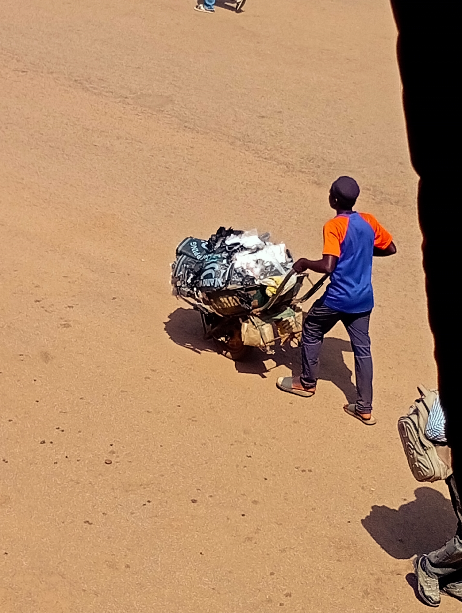 A person is pushing a cart loaded with items, likely merchandise, through an outdoor area with a dirt or sandy ground, characteristic of Bukuru, Nigeria. The person is wearing a blue t-shirt with orange sleeves and dark pants, along with sandals. The cart is laden with what appears to be clothing or textiles, some of which are white, black, and patterned. Visible text on the cart includes "SPPING" and "DUV". The scene suggests a daytime setting with warm weather, indicated by the bright sunlight and shadows. Another person, partially visible and wearing a backpack, is on the right side of the frame, also with their feet on the ground. The overall impression is of a person engaged in commerce or transportation of goods in a busy or frequented area.