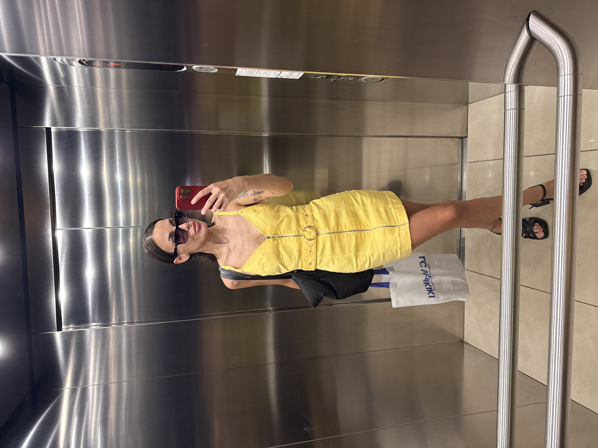 The image shows a woman taking a mirror selfie inside an elevator, rotated ninety degrees clockwise. The woman has dark hair and is wearing dark sunglasses, a yellow sleeveless mini-dress with a V-neckline, a front zipper, and a metallic ring detail at the waist. She has tattoos visible on her left arm, including a sun symbol. She holds a red smartphone, with its camera lens visible, to capture her reflection. She is wearing black flat sandals.
A black jacket or bag is casually hanging from her shoulder, and a white shopping bag with a blue logo is placed near her feet. The logo on the shopping bag clearly reads "LC WAIKIKI".
The elevator interior features brushed stainless steel walls, giving it a modern appearance. The floor is covered with light-colored, possibly beige or cream, tiles. A curved metal handrail is visible on the right side of the elevator, reflecting the surroundings. On the elevator control panel at the top, the number "2" and an upward-pointing arrow are illuminated, indicating the elevator's current floor and direction of travel. The woman's expression is partially obscured by her sunglasses, but her lips are slightly parted. The overall setting is consistent with an elevator in a commercial building, likely in Pleven, Bulgaria, given the provided context.