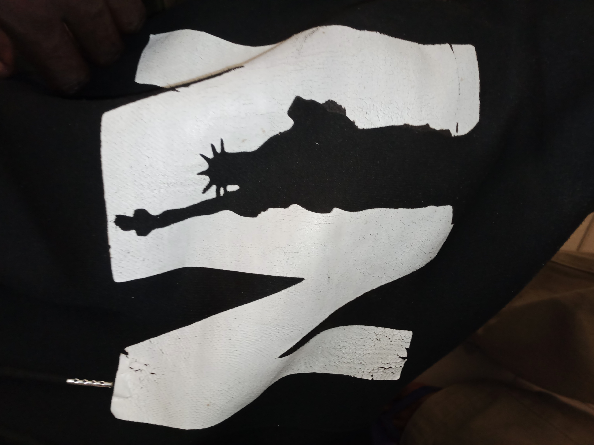 A black t-shirt with a white graphic of the Statue of Liberty is displayed. The graphic is a silhouette of the Statue of Liberty with its torch raised, set against a large, abstract white shape that resembles the number "2". The white graphic appears slightly worn and cracked in some areas. A human hand, with dark skin, is visible in the upper left corner, partially covering the t-shirt. The t-shirt is laid out on a dark surface, with hints of a person's legs and clothing in the background, suggesting it might be on a person's lap or a folded surface. The image is taken at close range, focusing on the graphic and the hand. There is no discernible information about the location in Jalingo, Nigeria.
