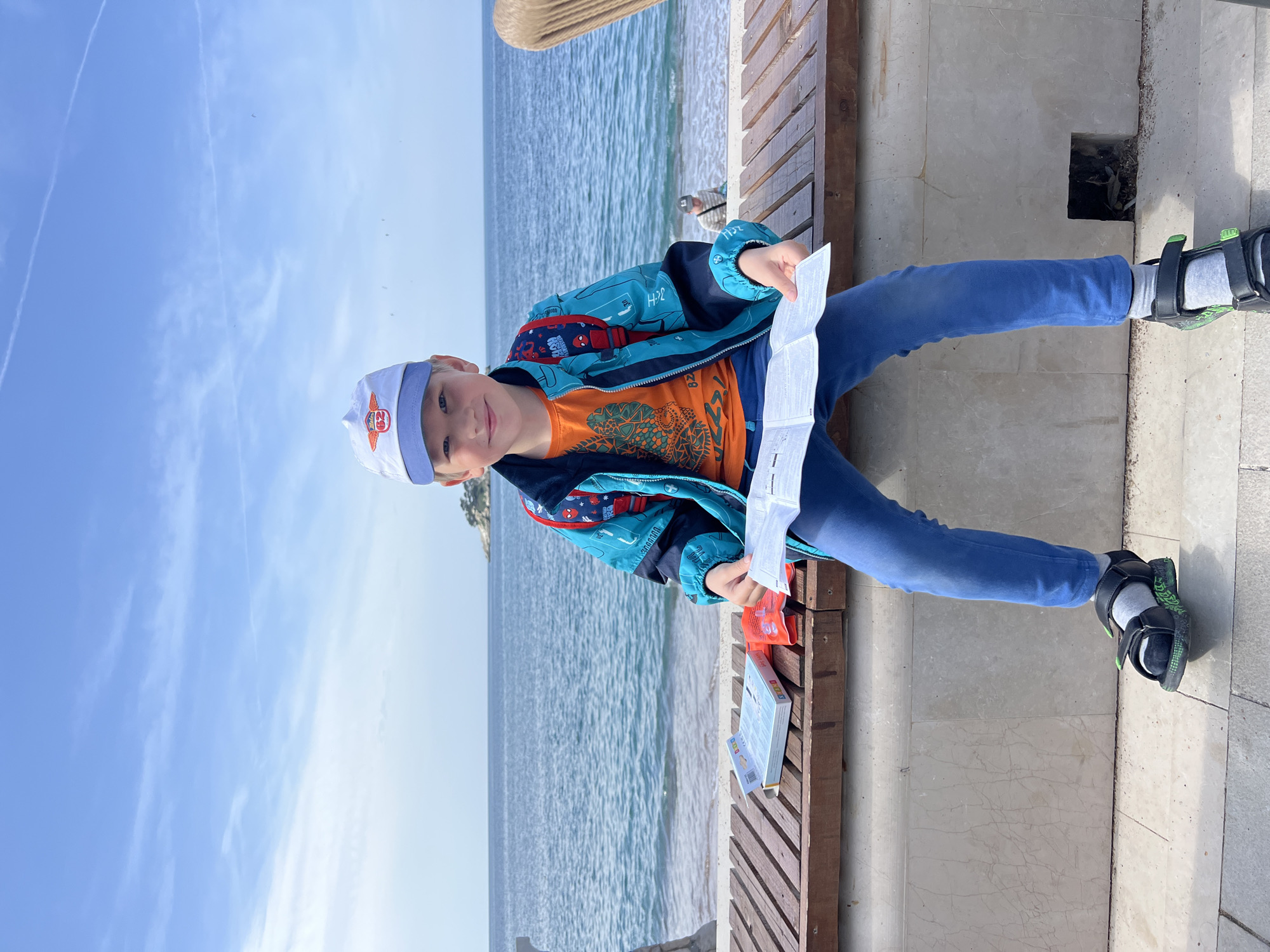 A young boy is sitting on a bench in Petrovac Centar, Montenegro. He is holding a map and appears to be studying it. He is wearing a light blue jacket, an orange t-shirt, blue pants, and black sandals. He also has a white cap on his head. The bench is made of wood and has a concrete base. In the background, there is a view of the sea and the sky, which is blue with some clouds. The location appears to be near the promenade, with a tiled surface in the foreground. The photo is oriented vertically.