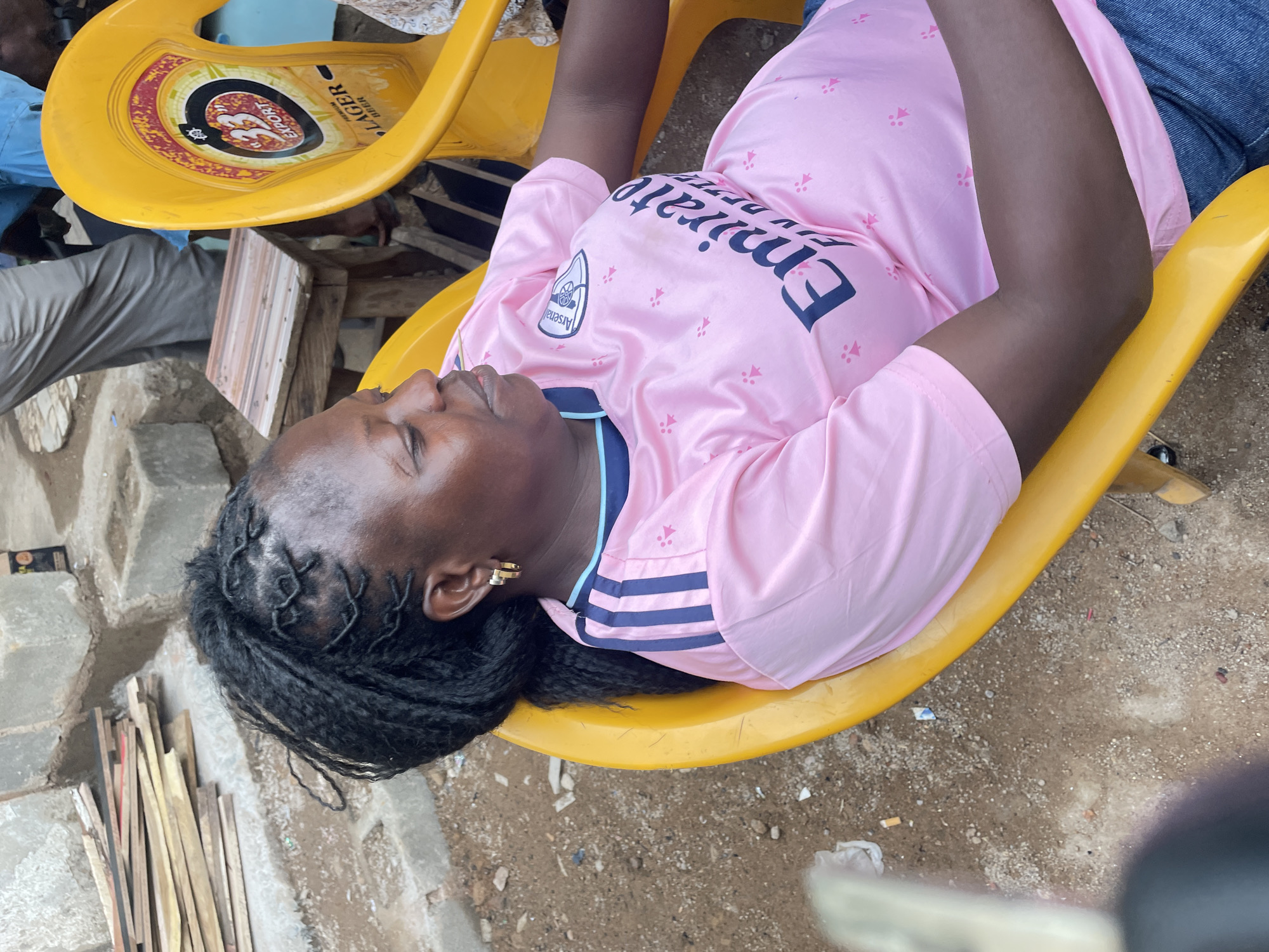 An overhead shot shows a woman lying on a yellow plastic chair in an outdoor environment in Jos, Nigeria. The woman wears a pink Arsenal soccer jersey. Her hair is styled in black braids. She wears gold hoop earrings. The chair is situated on a patch of dirt and gravel. In the background, there are stacks of wood, another yellow plastic chair with a beer logo, and legs of other people.