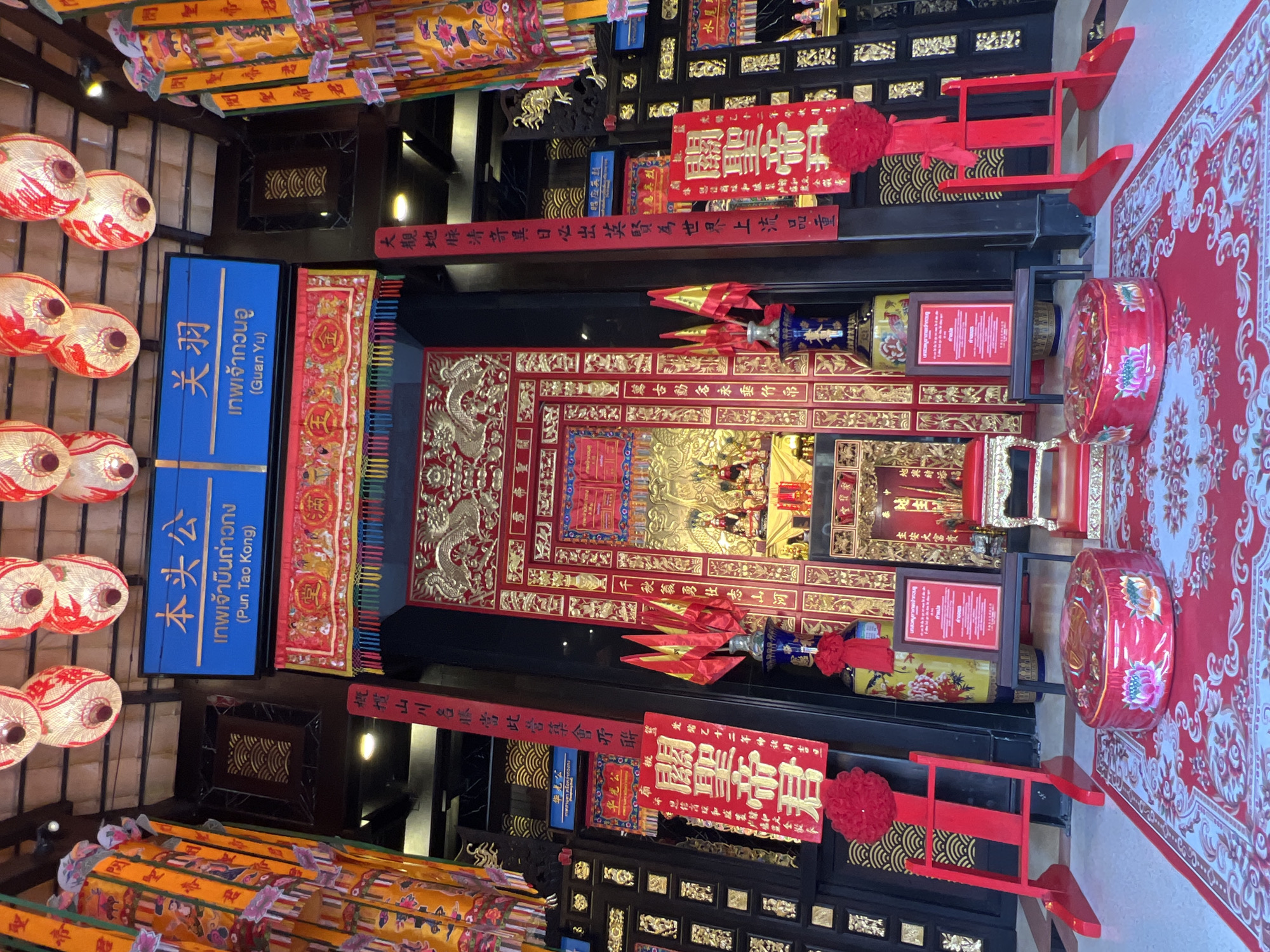 The image depicts the interior of a brightly decorated temple in Baan Hua Thanon, Thailand. Dominating the scene is an ornate, gold and red altar with a red carpet leading up to it. The altar features various religious statues and offerings. Flanking the altar are large vases and red banners with Chinese characters. Overhead, numerous decorative lanterns hang from the ceiling. The walls are adorned with intricate gold patterns. In the foreground, a red patterned rug covers the floor, with decorative red cushions placed on it. The overall atmosphere is one of vibrant religious and cultural display.