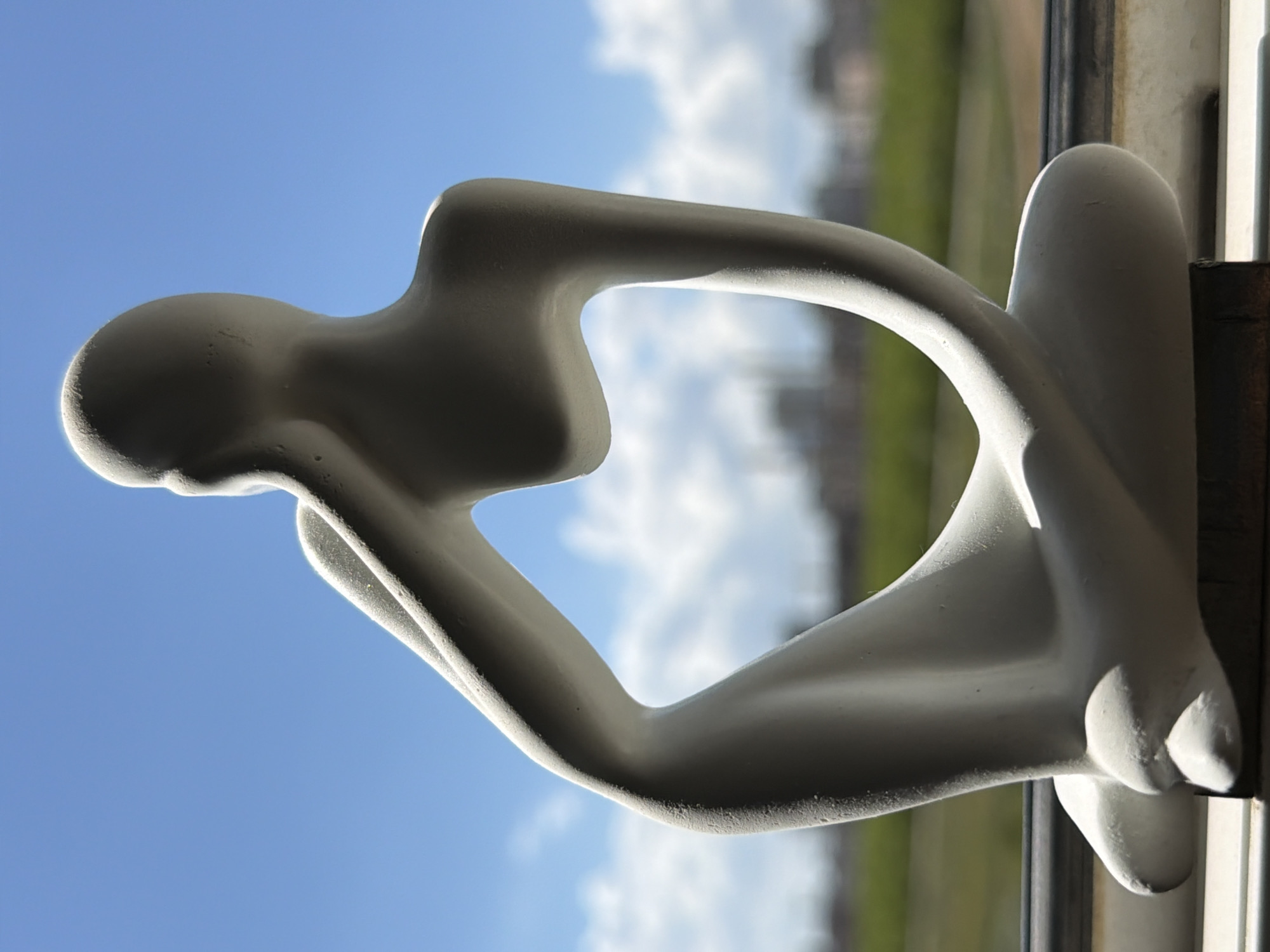 A white, abstract sculpture of a seated, pensive human figure is positioned on a windowsill. The sculpture is made of a matte material and depicts a figure with its hand resting on its head. The background shows a clear blue sky with scattered white clouds, and a blurred green landscape with distant buildings. The image does not contain any information that would suggest it is located in Astana, Kazakhstan.