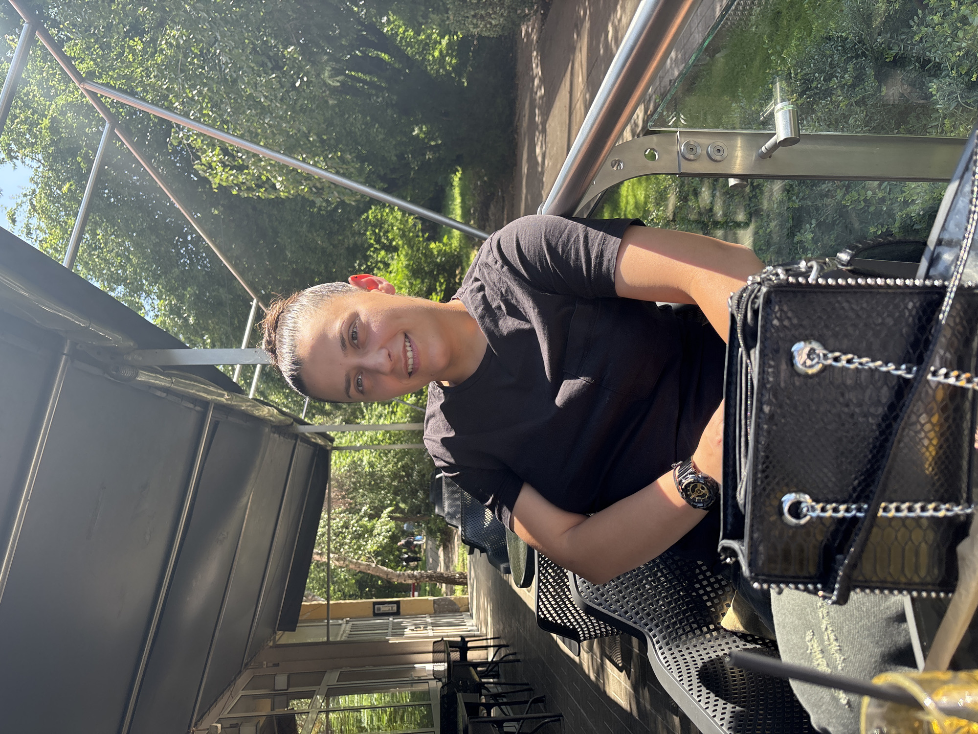 A person is seated outdoors at a cafe or restaurant patio, captured in a portrait-style shot. The individual, seen from the waist up, is smiling and looking directly at the camera. They have dark hair tied back, light skin, and are wearing a black short-sleeved t-shirt and dark pants, possibly jeans with distressed areas. A black wristwatch with gold accents is visible on their left wrist.
To their right, a black handbag with a patterned texture and silver chain straps rests on the table, partially obscuring a drink with a straw. The person is sitting on a modern perforated metal chair, which also makes up the table surface.
The setting appears to be an elevated or balcony-style outdoor area, with a metal railing and a glass barrier separating the patio from the surroundings. Beyond the barrier, lush green trees and foliage are abundant, suggesting a park or garden area nearby. A paved pathway can be seen through the trees. In the background to the left, there's a building facade with glass windows and outdoor seating areas with empty tables and chairs. The scene is brightly lit by natural sunlight, casting visible shadows and indicating it's daytime, likely midday or afternoon given the strong light. The overall mood appears pleasant and relaxed. No legible text is visible in the image. The location is Sofia, Bulgaria.